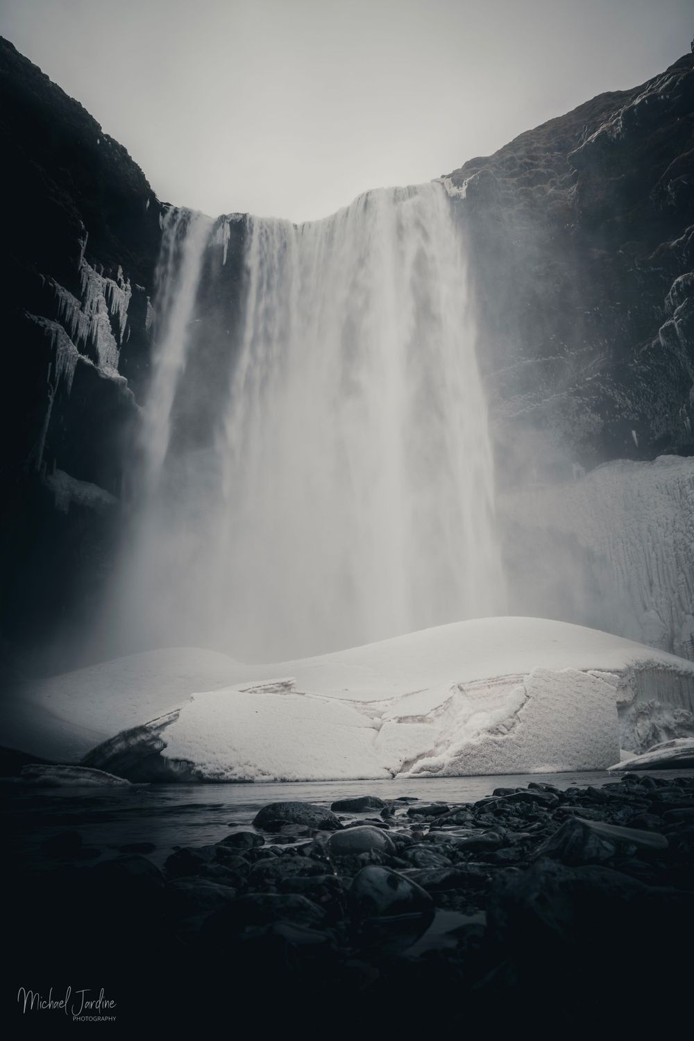 Powerful waterfall cascading down dark cliffs into snowy landscape below.