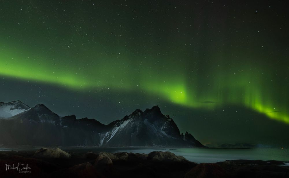 Green aurora borealis dancing over dark mountain peaks against starry night sky.