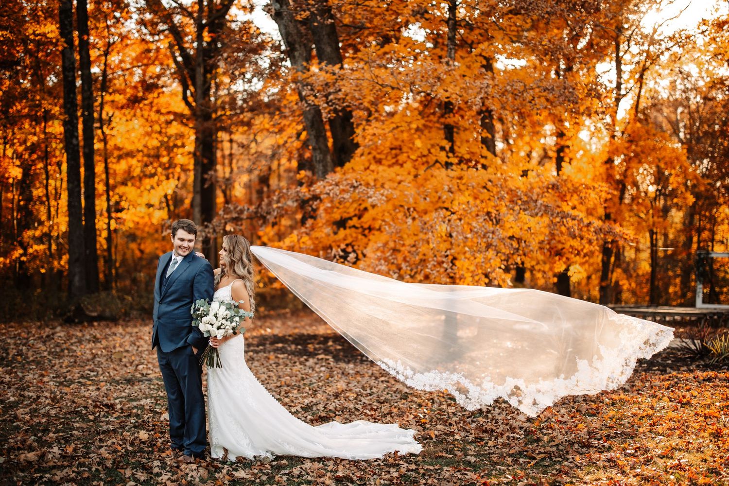 Wedding couple in autumn forest with bride's veil dramatically flowing in orange foliage.