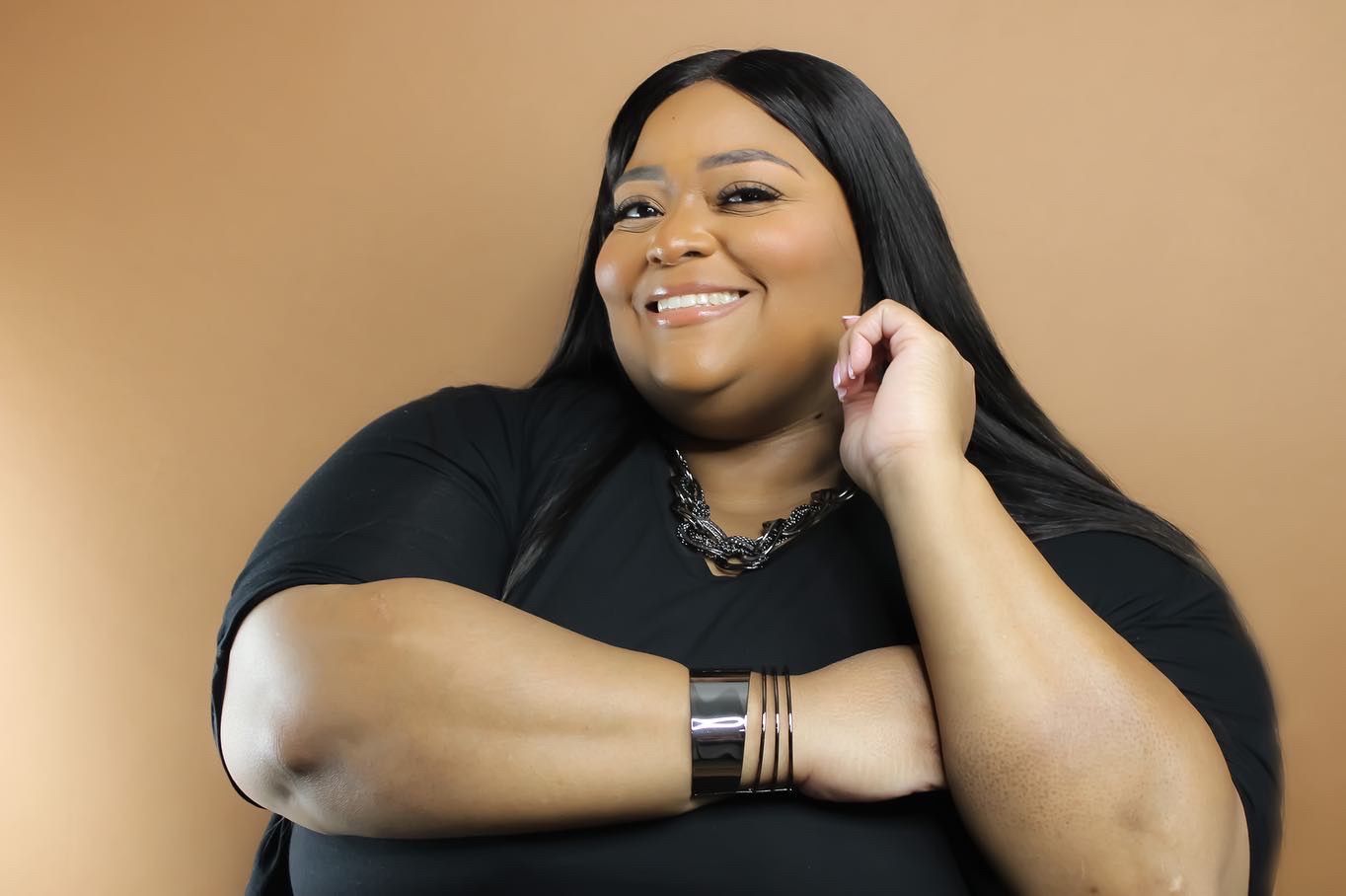 A person in a black top wearing multiple bracelets poses against a warm brown background.