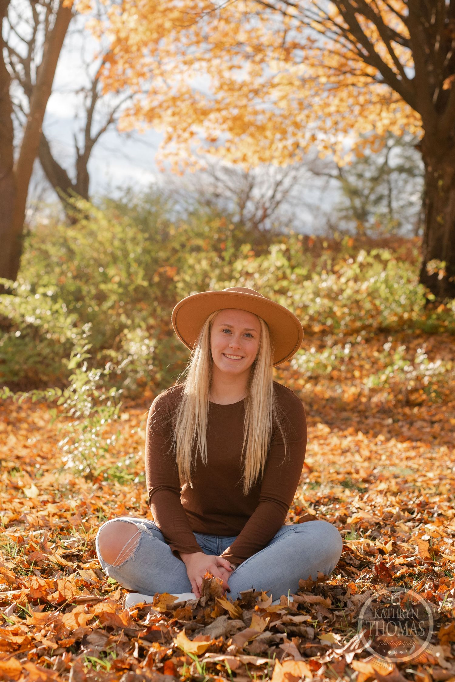 Person sits cross-legged on autumn leaves wearing brown sweater and wide-brimmed hat.