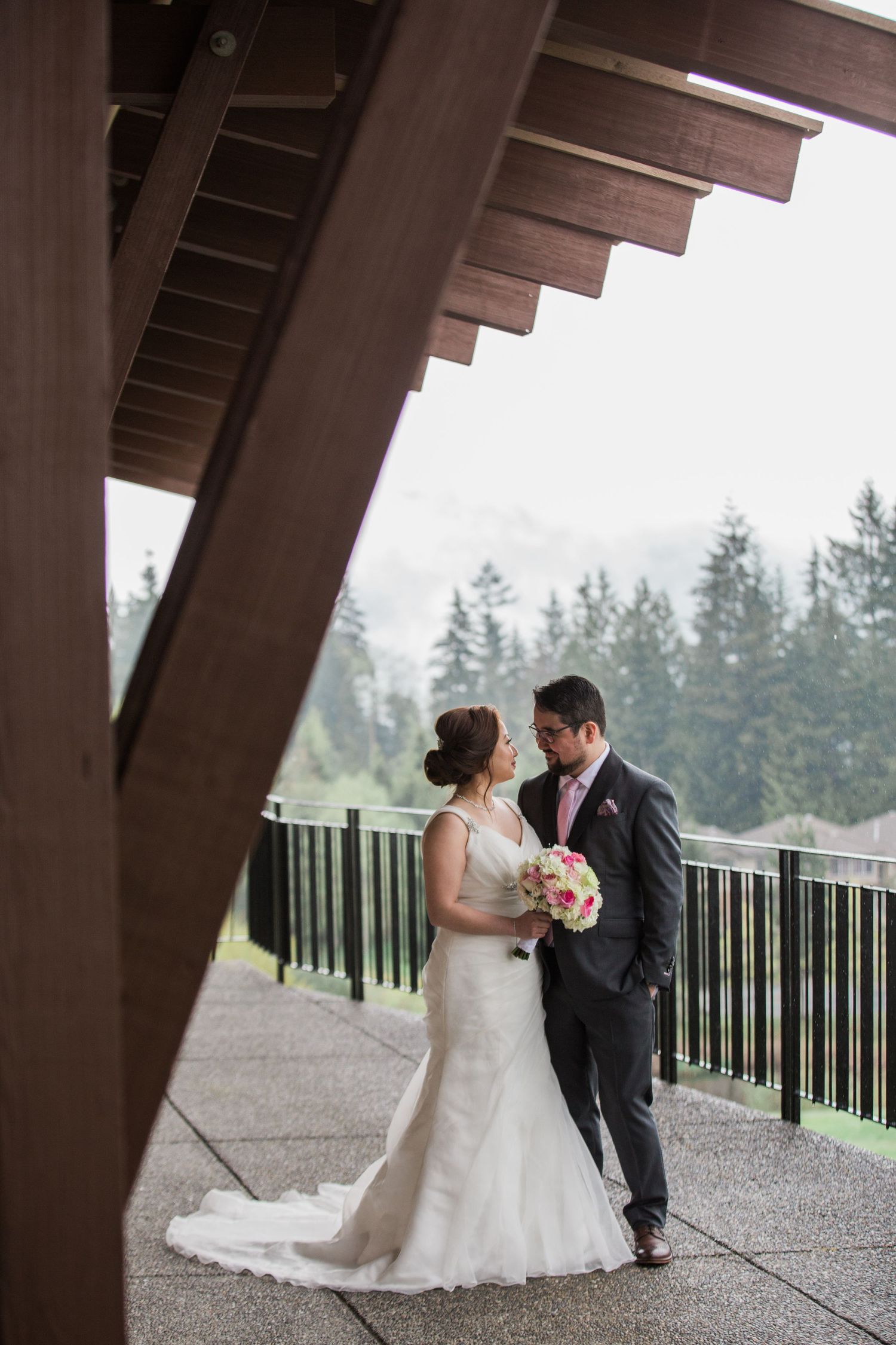 Wedding couple standing under wooden beams on covered outdoor terrace overlooking forest.