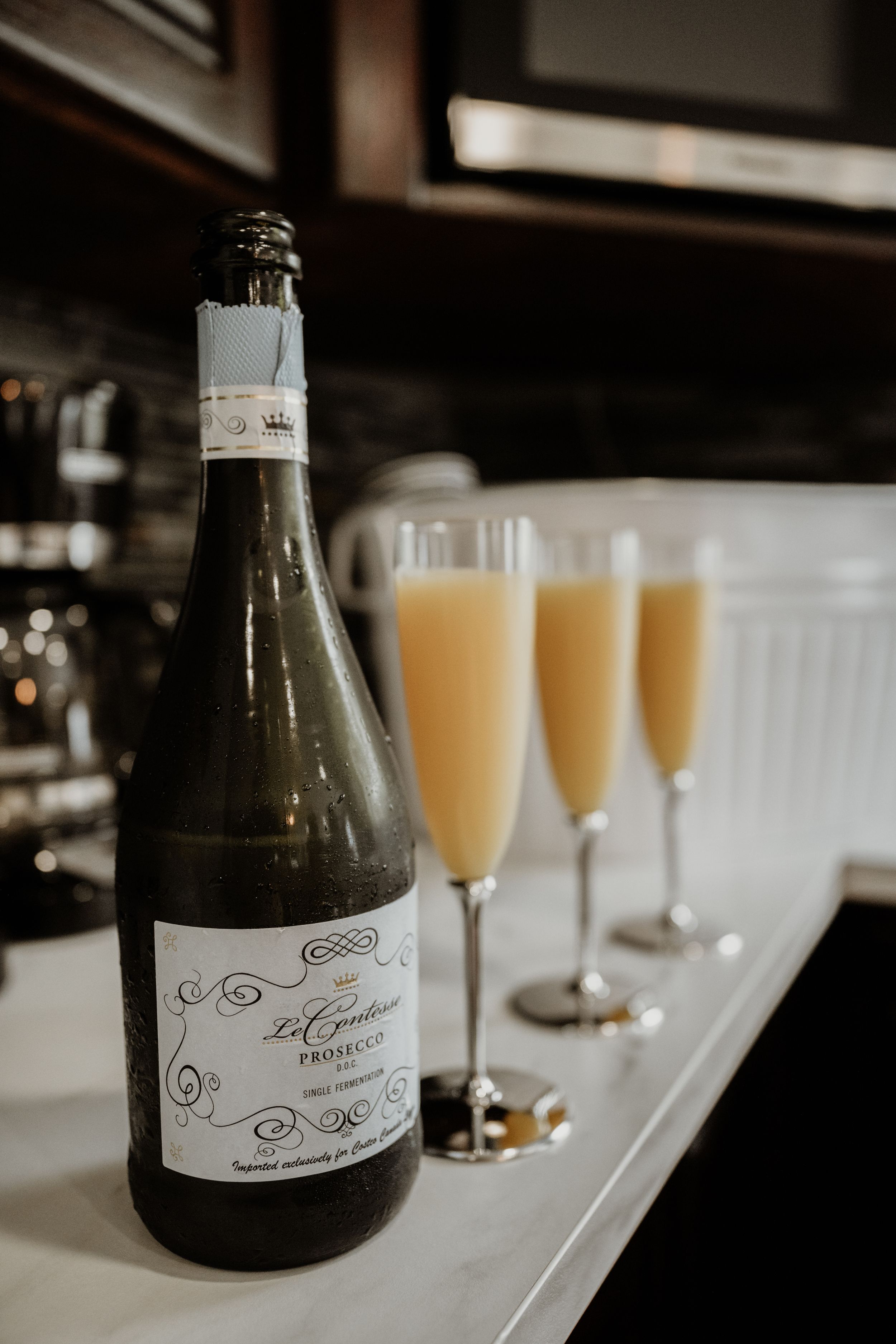 A bottle of champagne with three filled mimosa glasses lined up on a white counter at a bar.