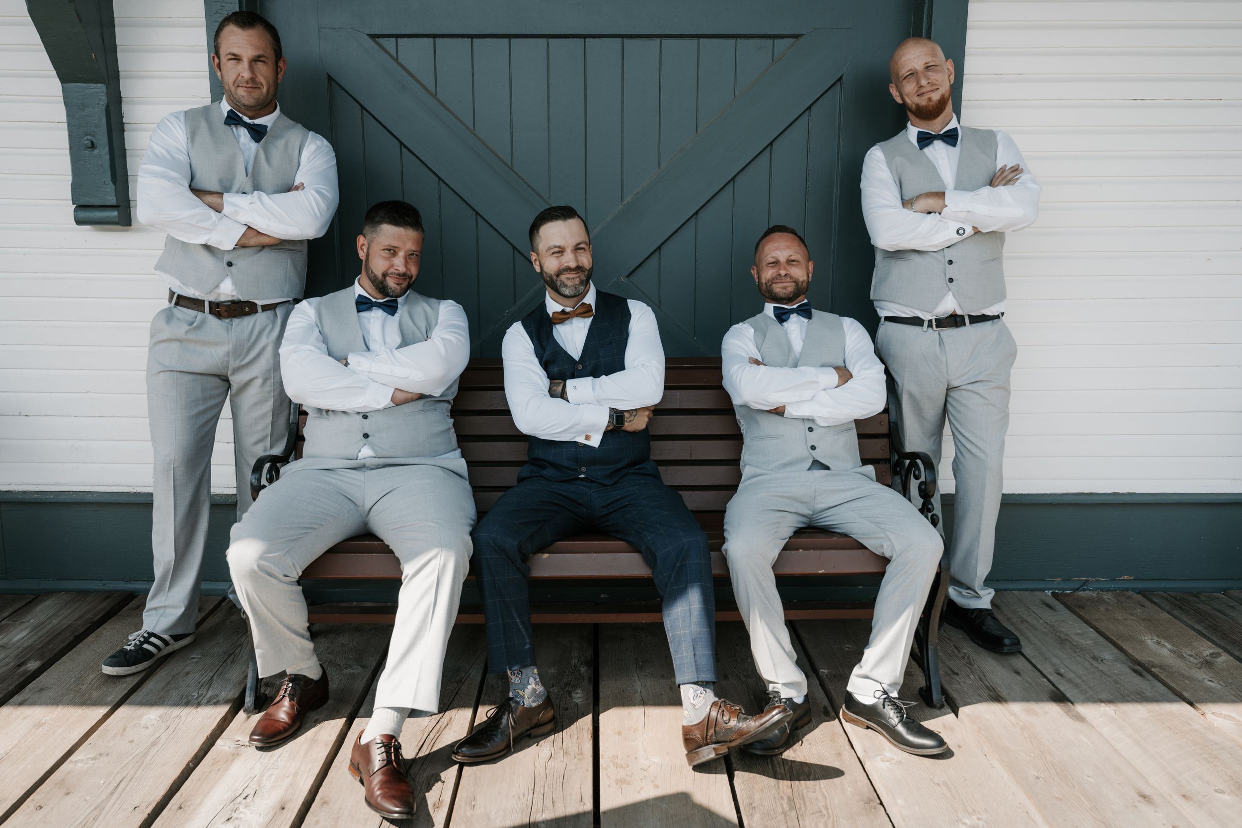 Wedding party in matching light gray suits sitting together on wooden stairs against a barn door background.