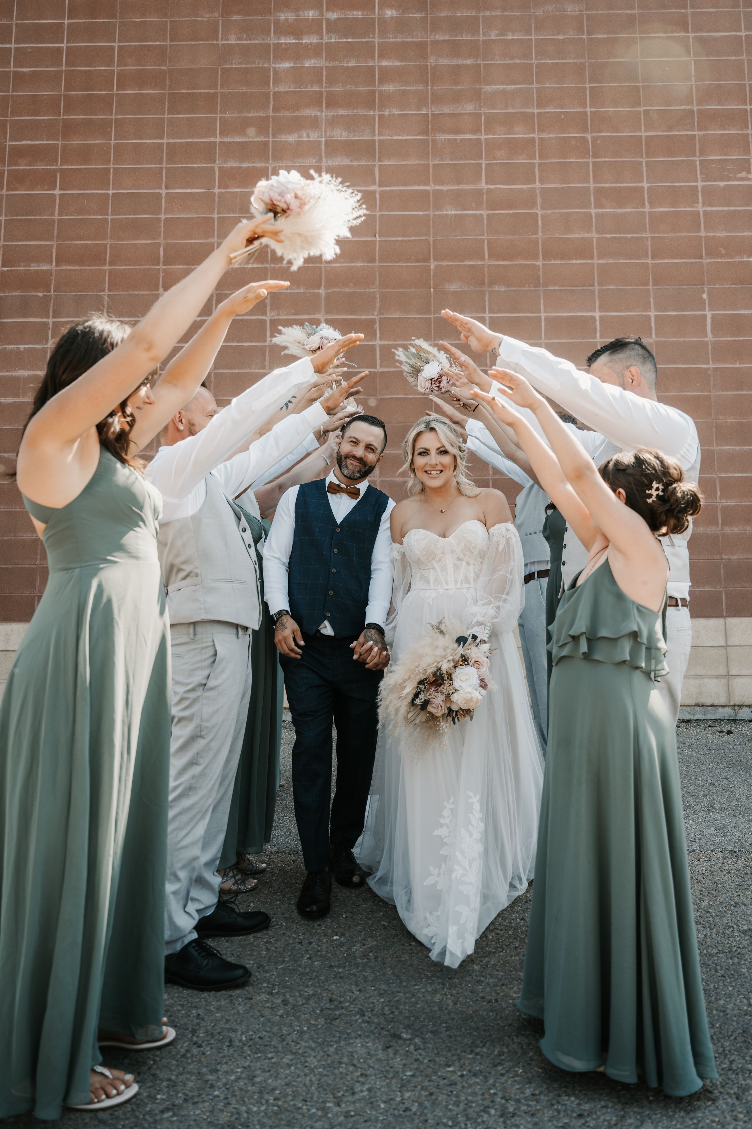 Wedding party members in sage green dresses toss confetti over newlyweds against a brick wall backdrop.