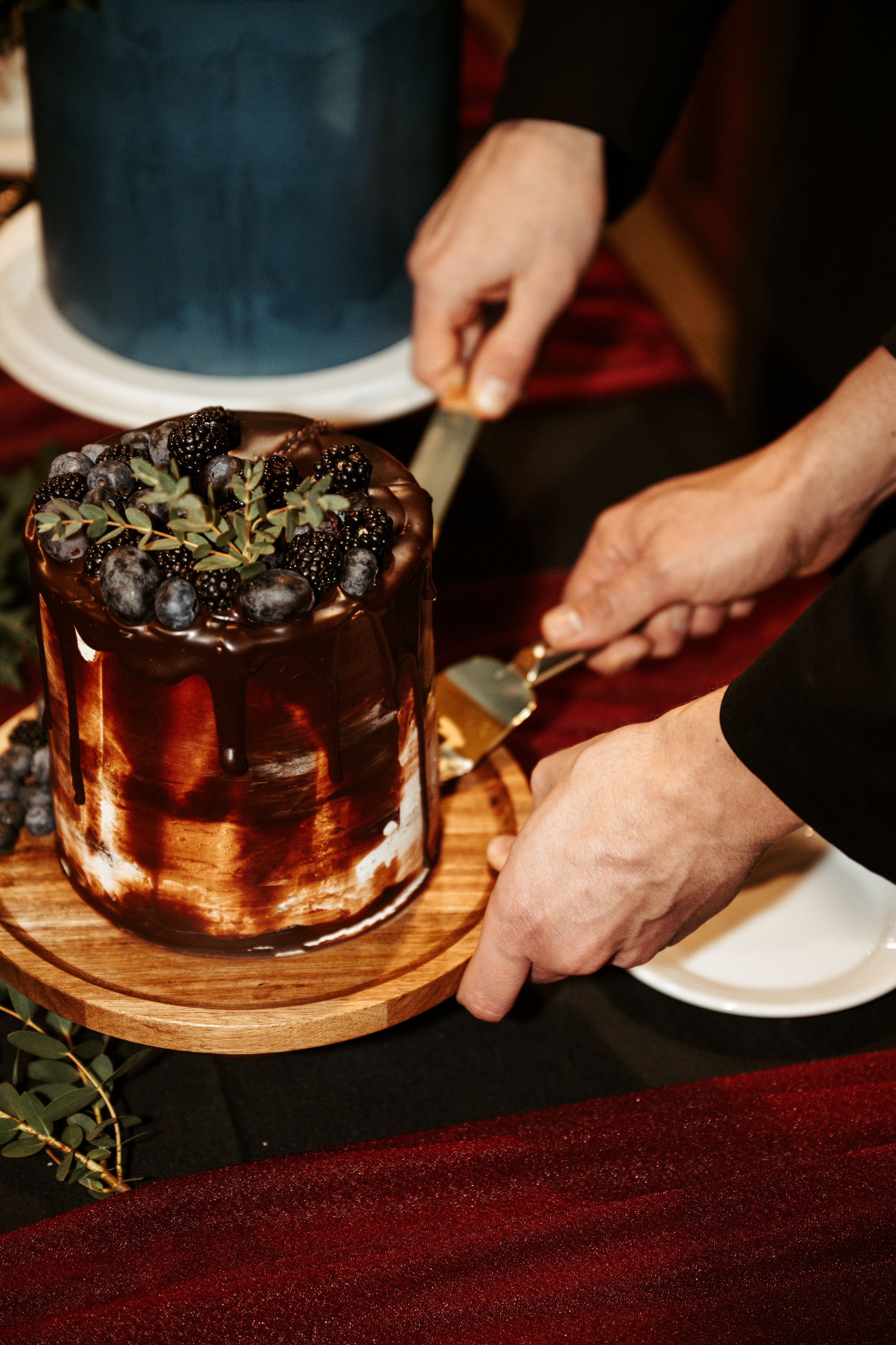 A decadent chocolate cake topped with fresh grapes sits on a wooden serving board as hands prepare to serve it.