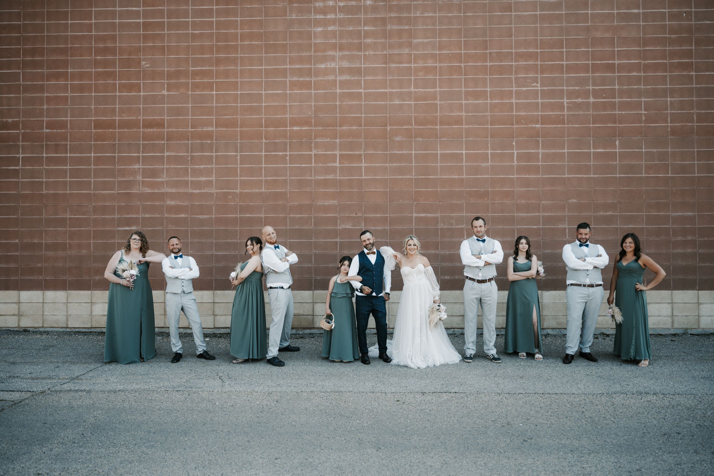 Wedding party in sage green attire poses against brick wall at modern celebration.