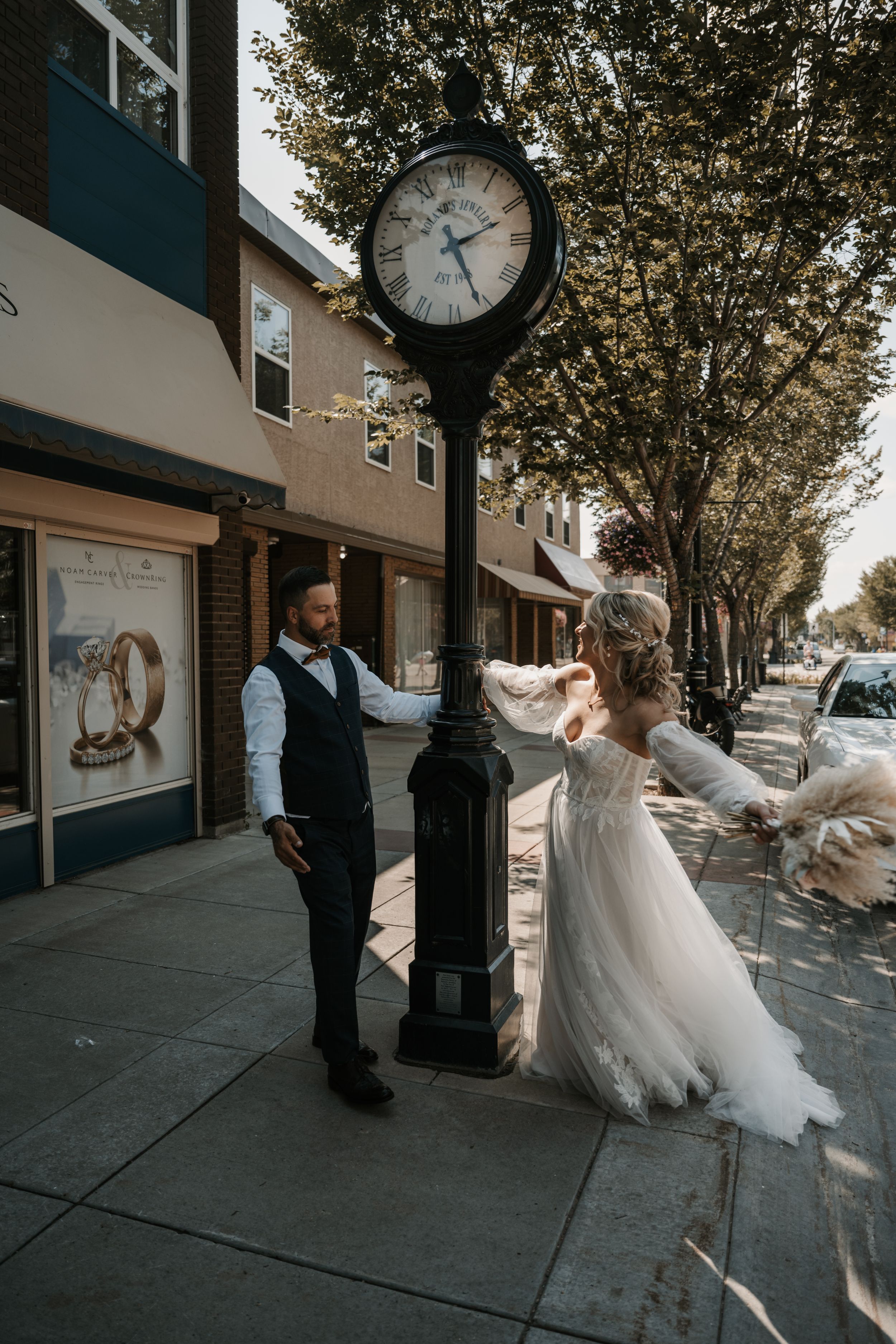 A couple in wedding attire dance playfully by a street clock on a tree-lined city sidewalk.