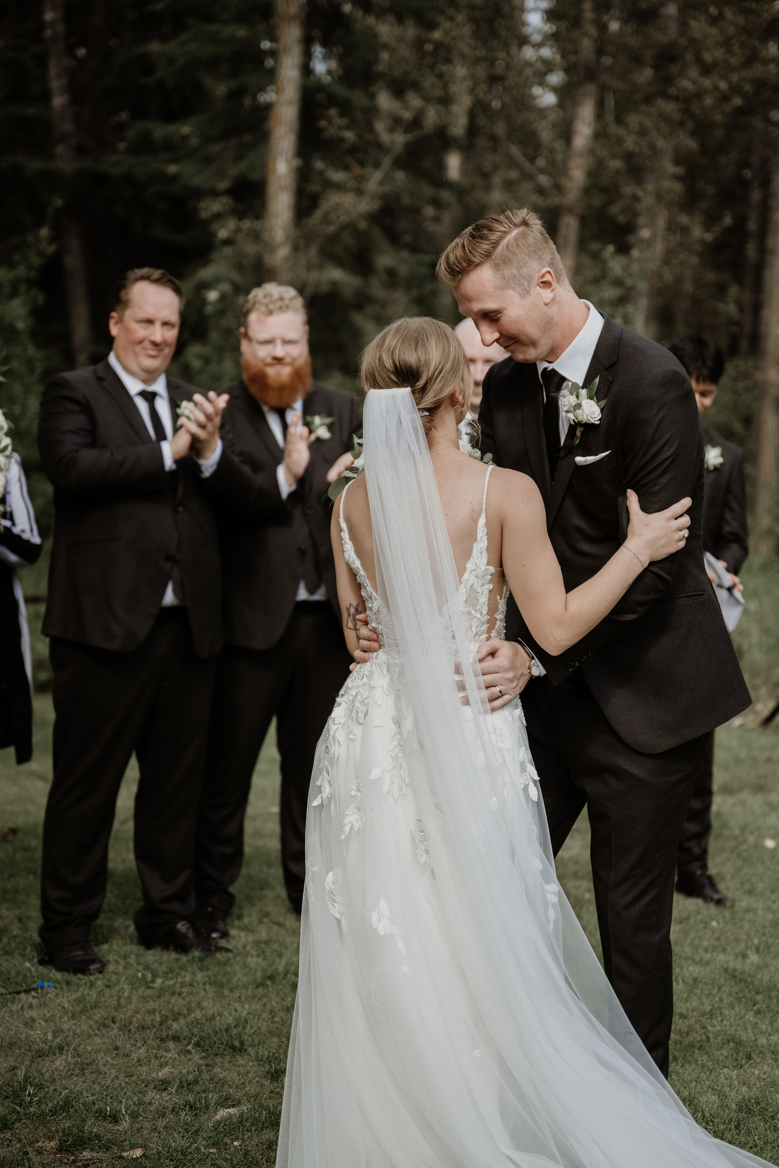 Newlyweds share intimate moment at outdoor wedding ceremony while guests applaud in forested background.
