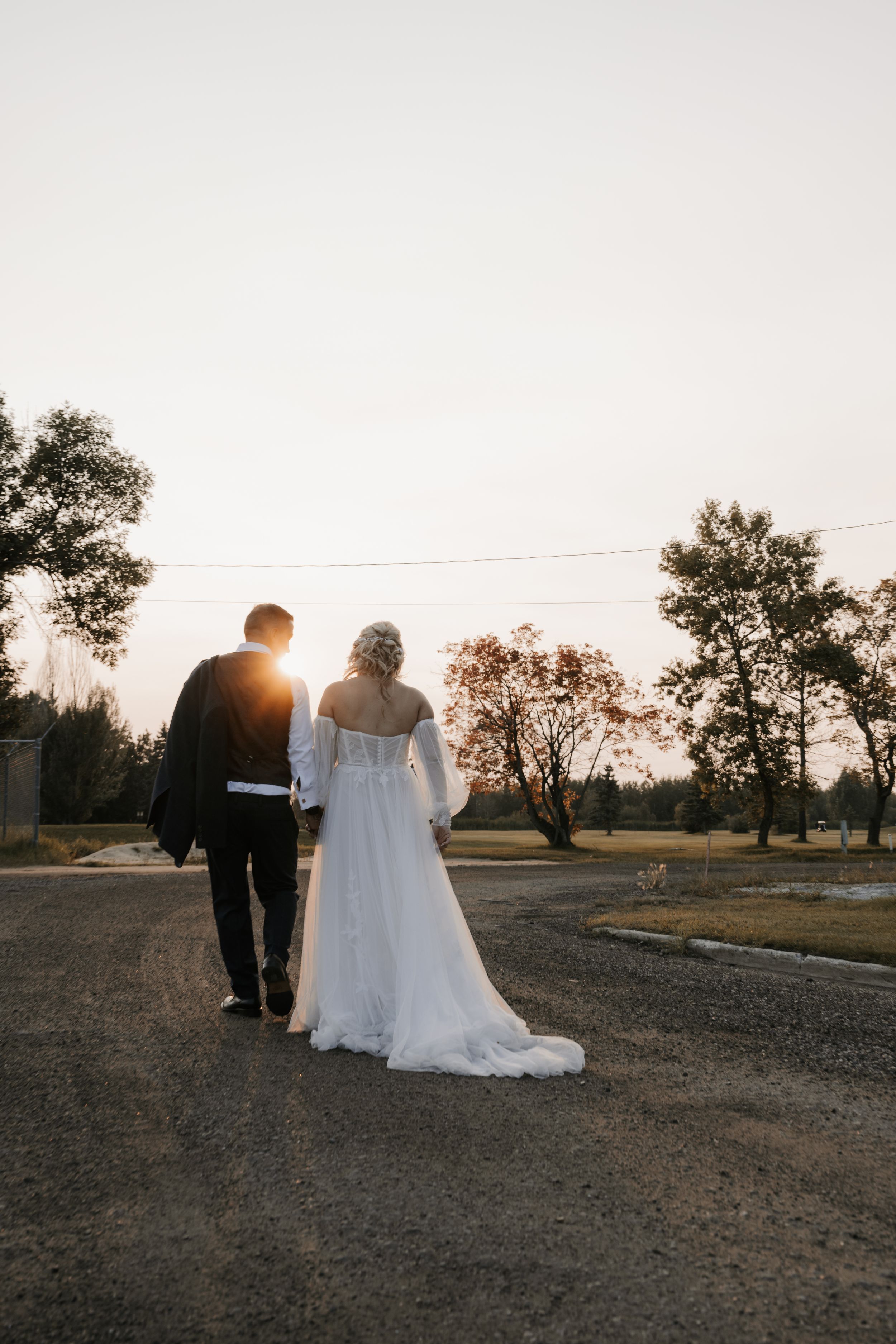 Couple in wedding attire walking down a dirt road at sunset with trees silhouetted against the sky.