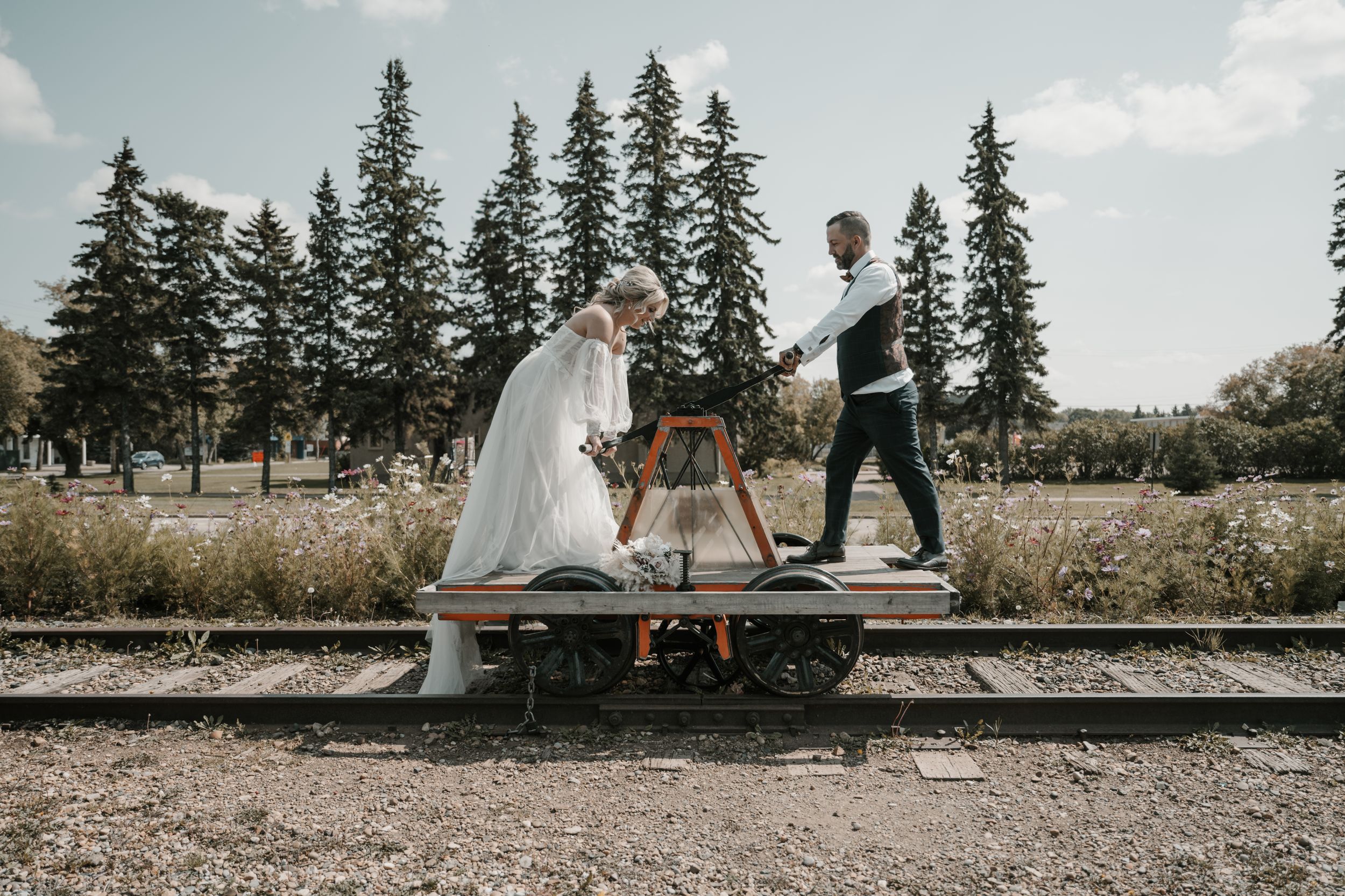 Three people stand on railroad tracks in a scenic forest setting with tall evergreen trees in the background.