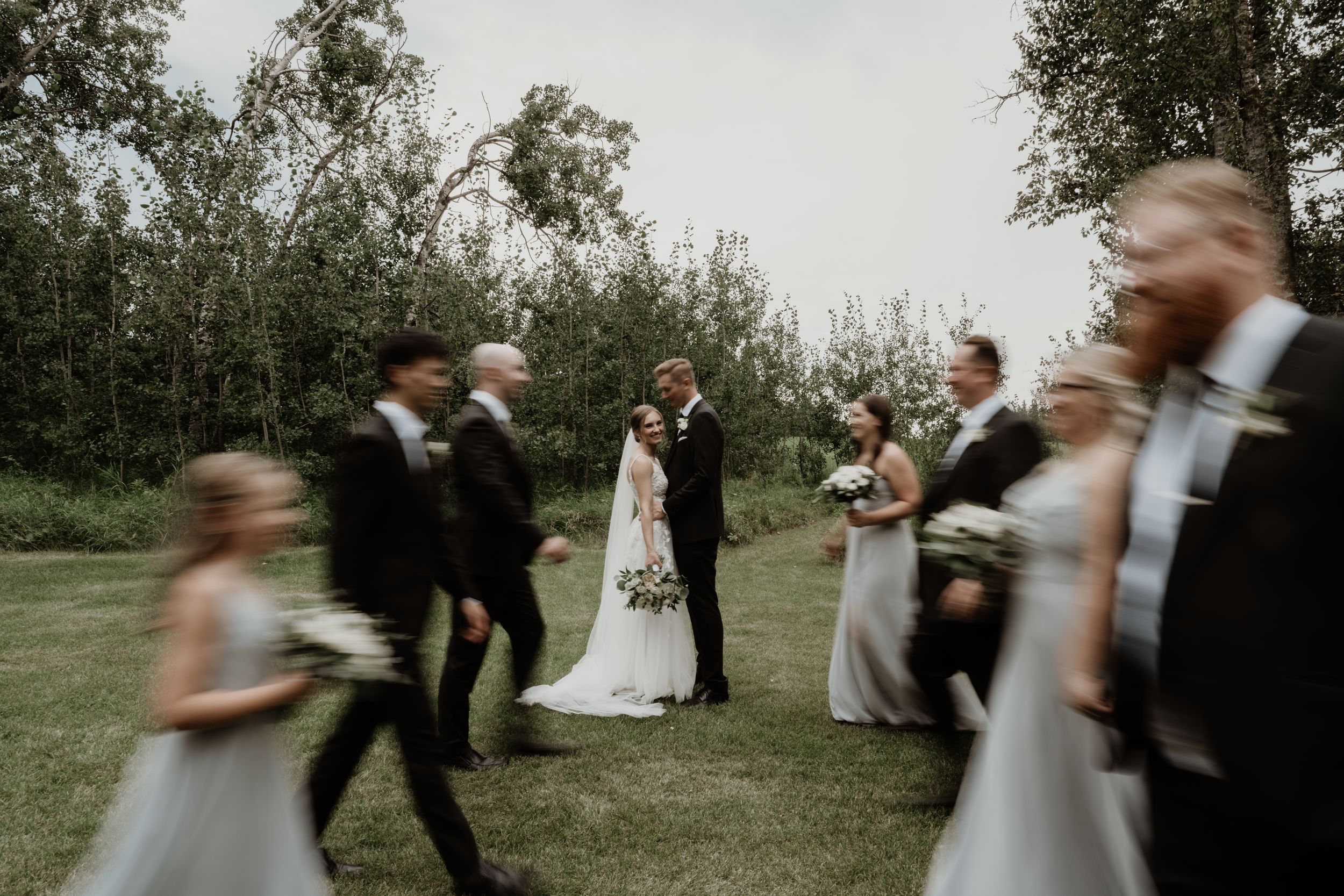 Motion-blurred wedding party celebrates outdoor with bridal party members lined up on grassy field surrounded by trees.