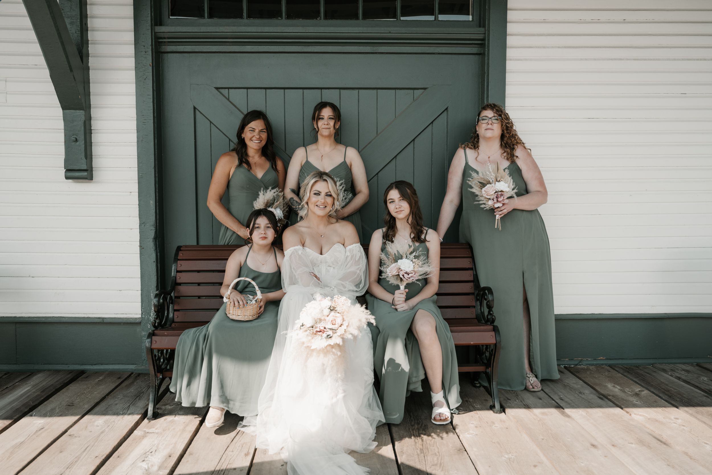 Wedding party in sage green and neutral dresses pose together on wooden porch steps against white building.