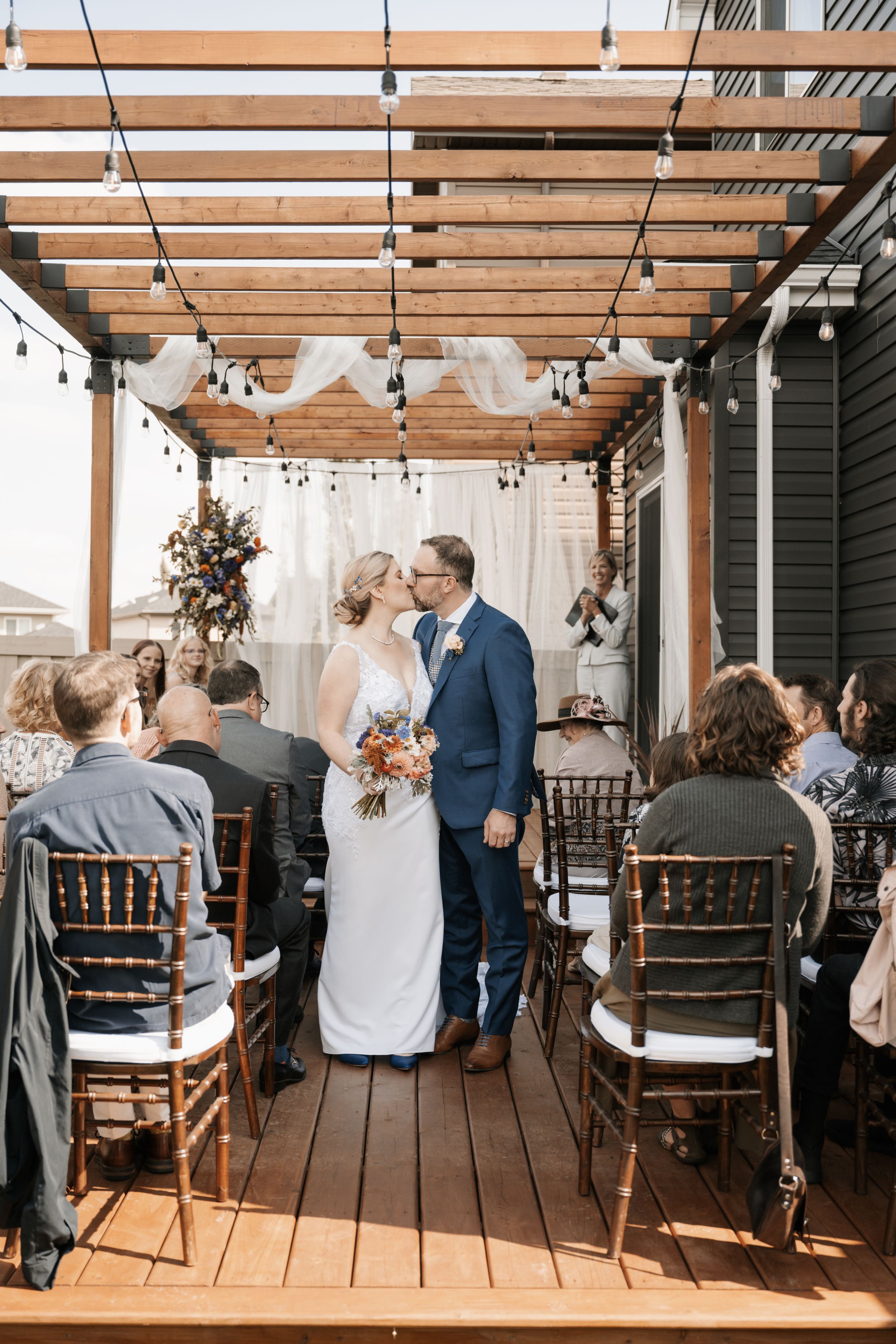 Intimate outdoor wedding ceremony under wooden pergola with string lights and white fabric draping on rustic deck.