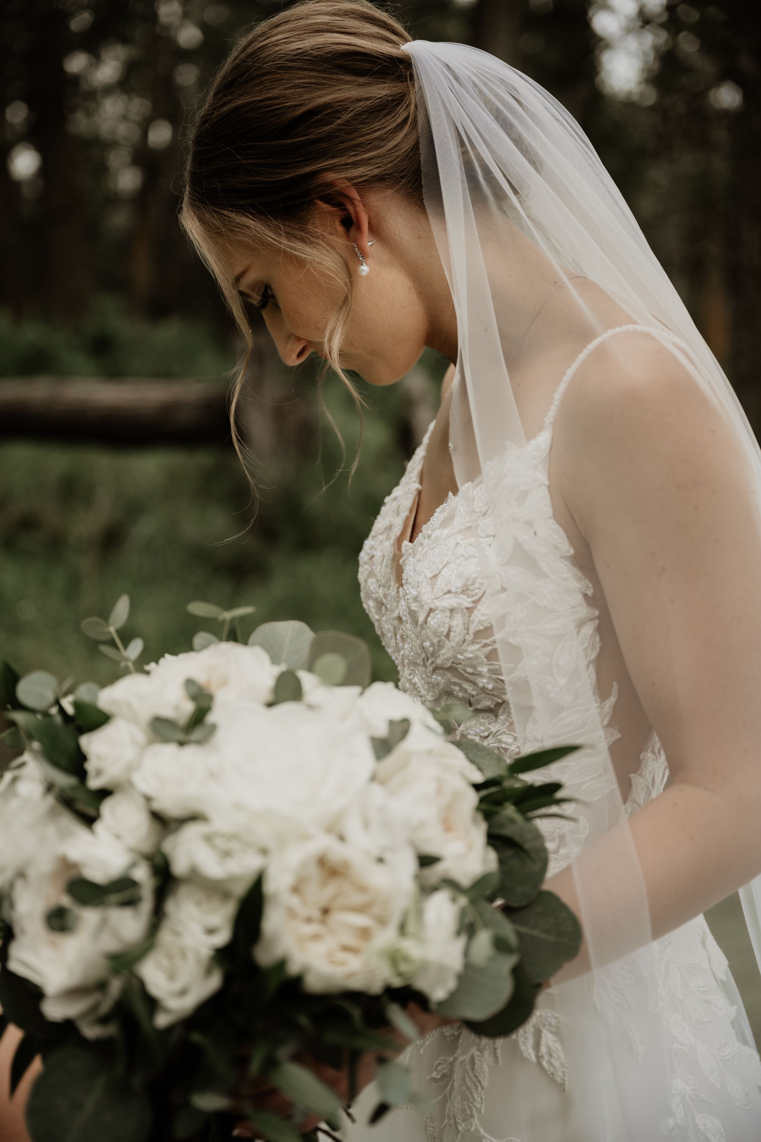 A bride in a lace wedding dress looks down at her elegant white bouquet with greenery in a moody outdoor setting.