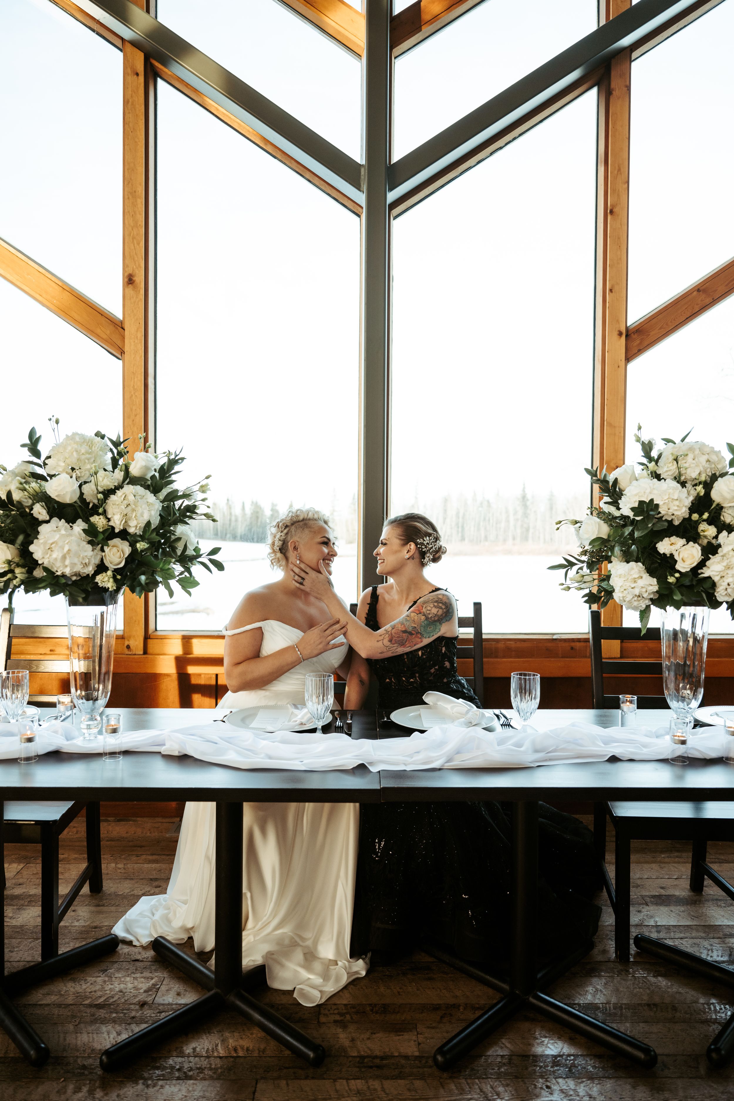 Two figures in contrasting black and white gowns share an intimate moment at an elegant reception table with floral arrangements.