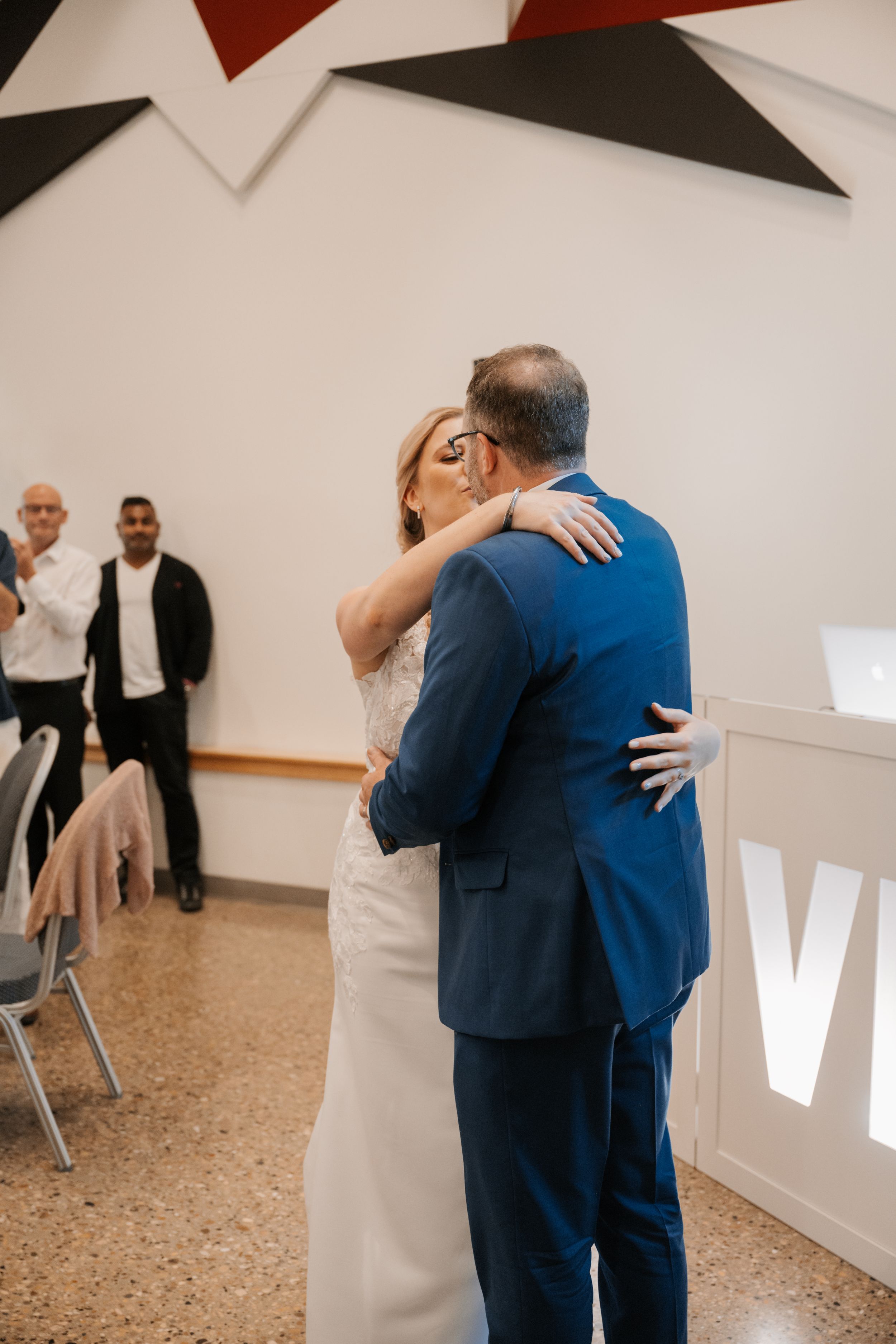 A couple embraces during their first dance at a wedding reception in a modern venue with white walls and geometric decor.