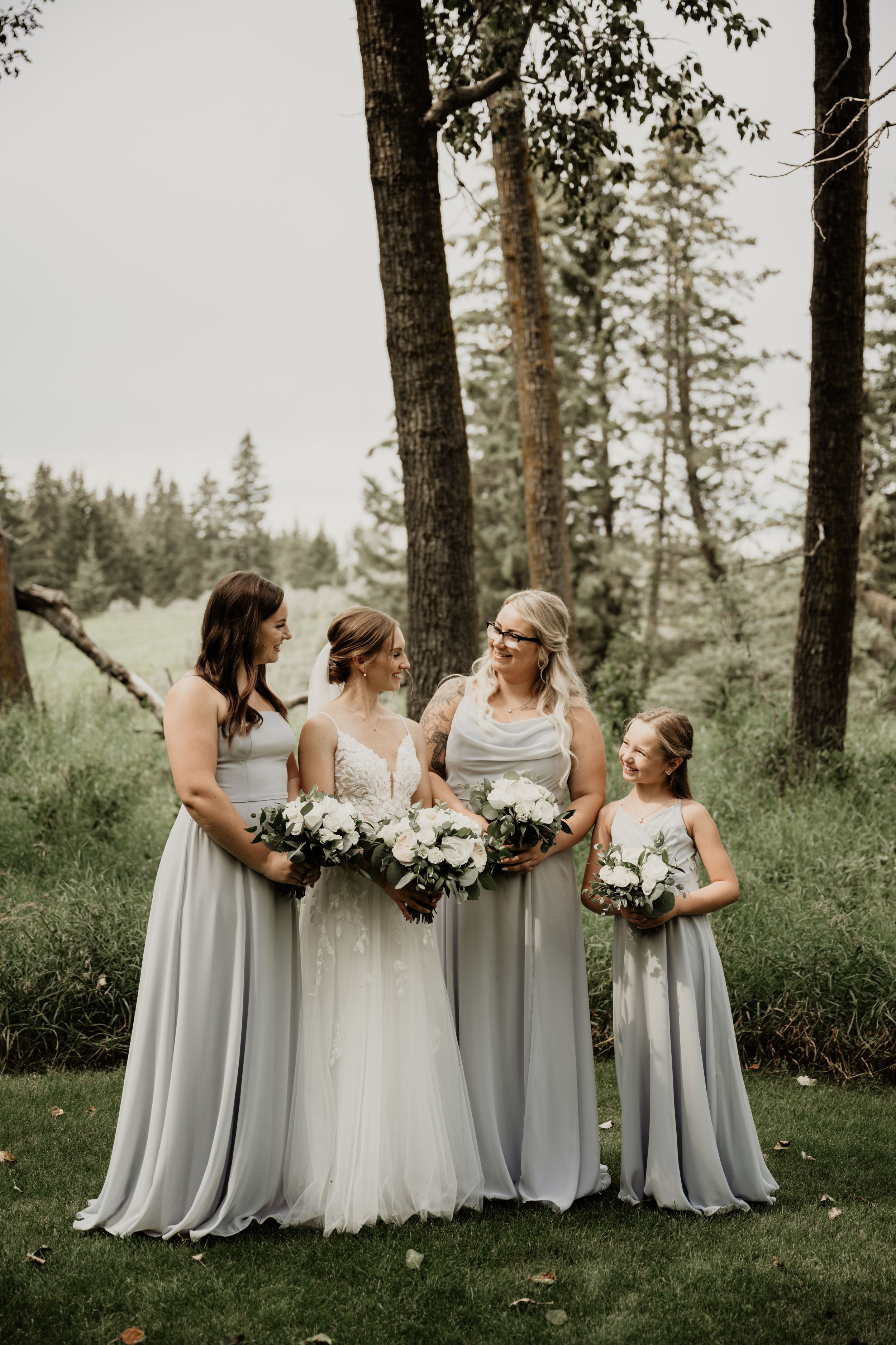 A bridal party in light gray dresses poses on a grassy hillside with white floral bouquets among tall evergreen trees.