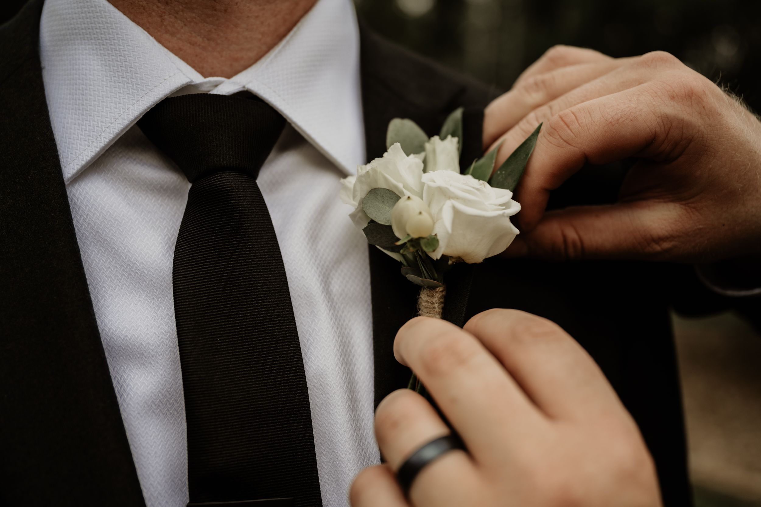 Someone helping to pin a white rose boutonniere on a black suit with white shirt and black tie during a formal event.