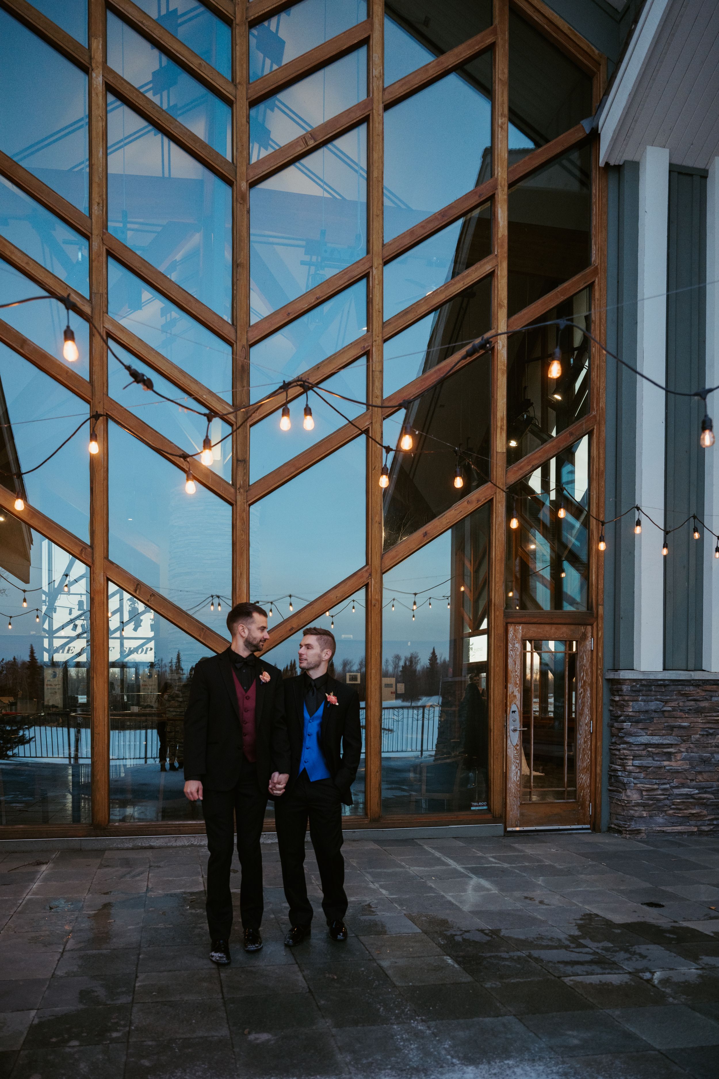 A couple stand together in front of a modern building with dramatic geometric windows and string lights at dusk.