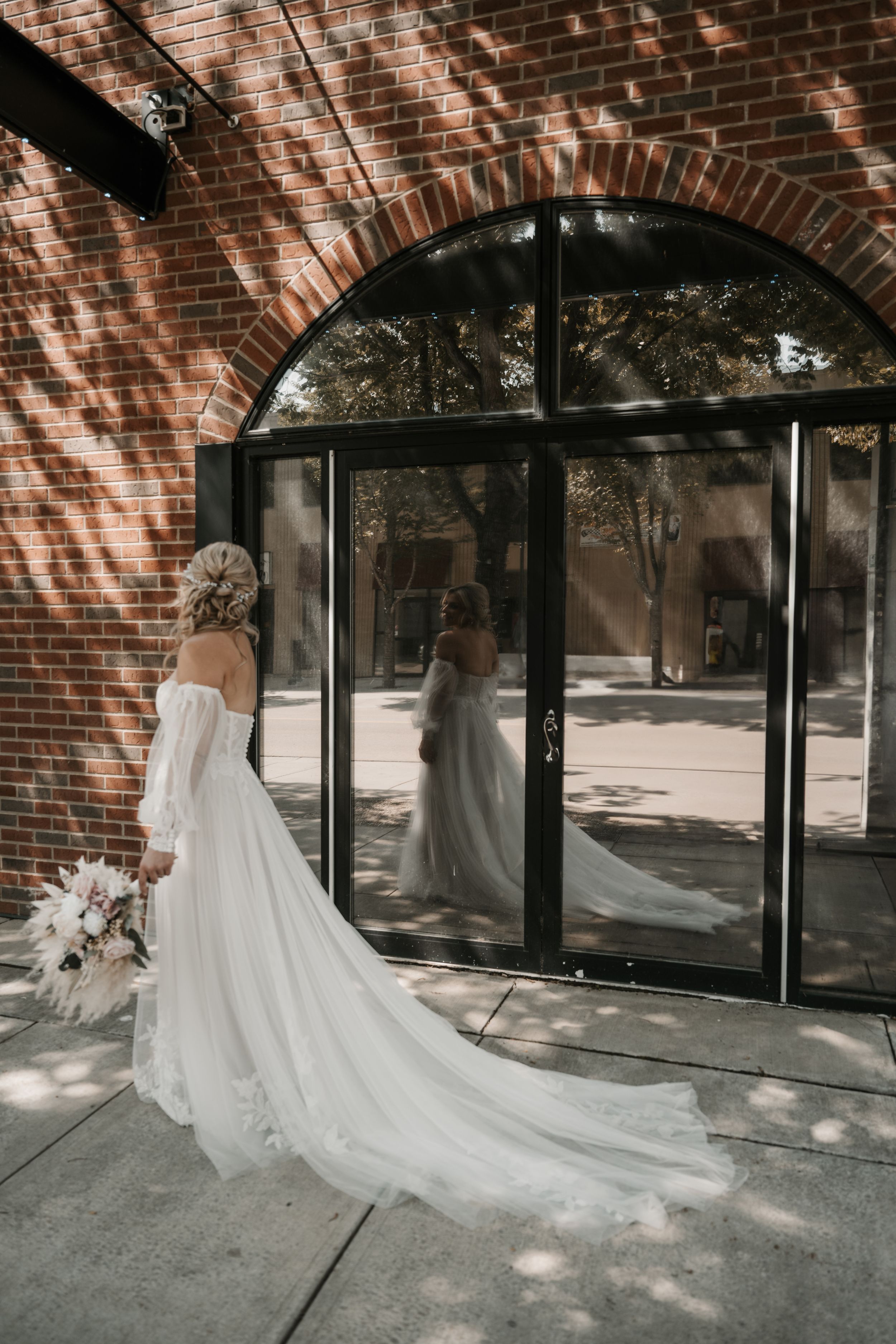 A person in a flowing white wedding dress with a long train stands before glass doors of a brick building.