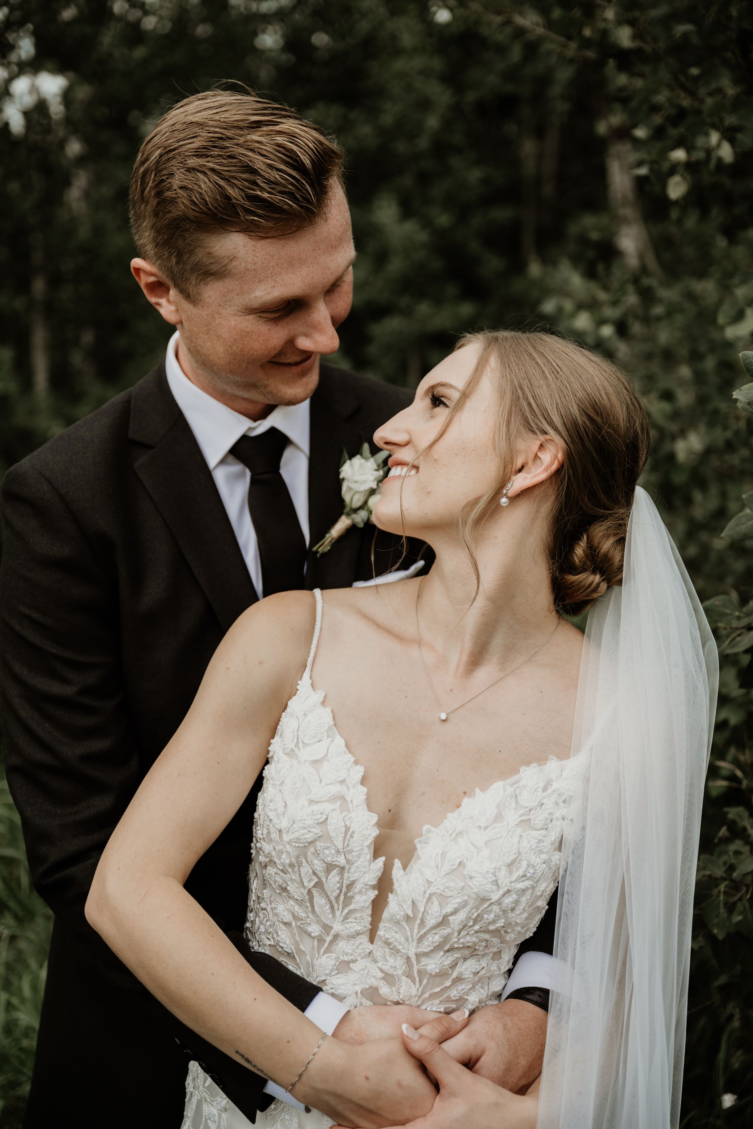 A bride in a lace wedding dress with veil shares an intimate moment with her groom in a dark suit amid greenery.