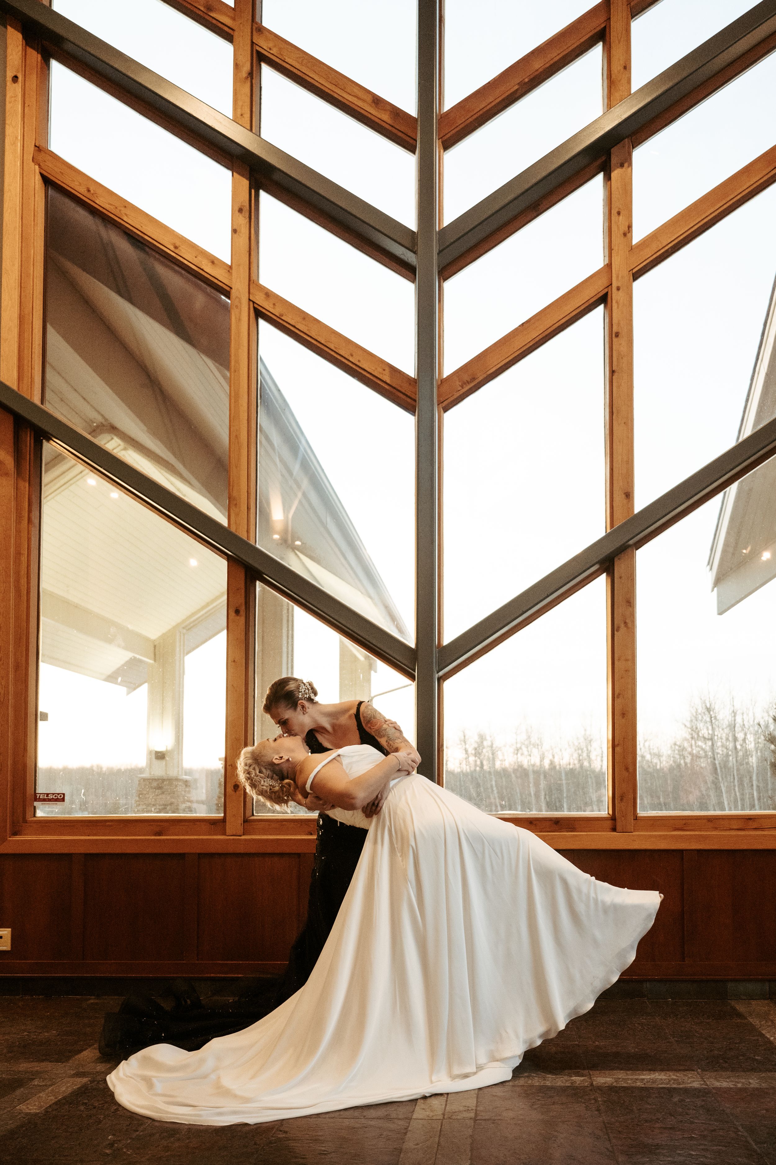 A romantic wedding photograph captured in front of tall wooden framed windows with natural light streaming through.