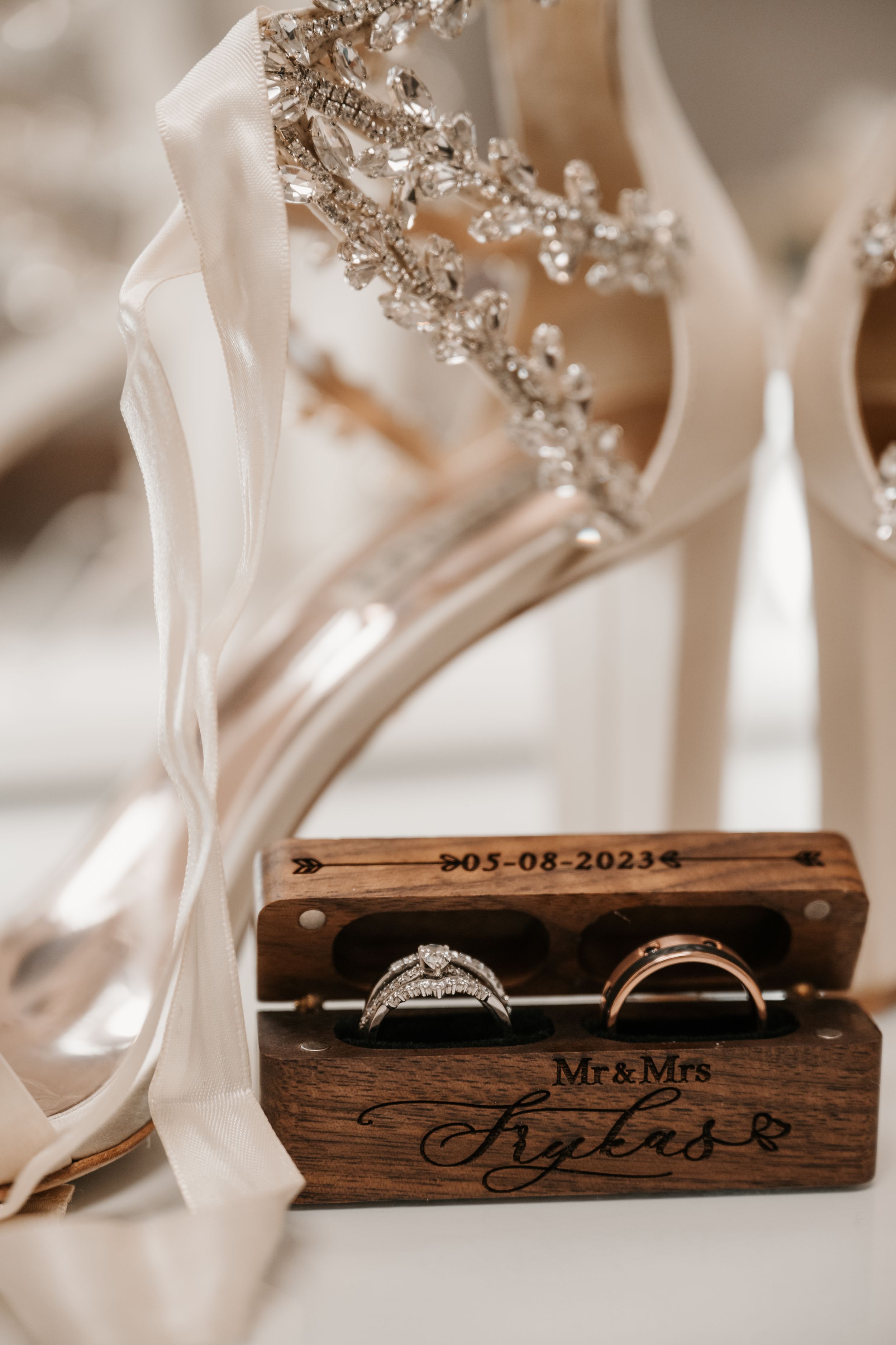 Wedding rings displayed in a personalized wooden box alongside bridal heels with crystal embellishments.