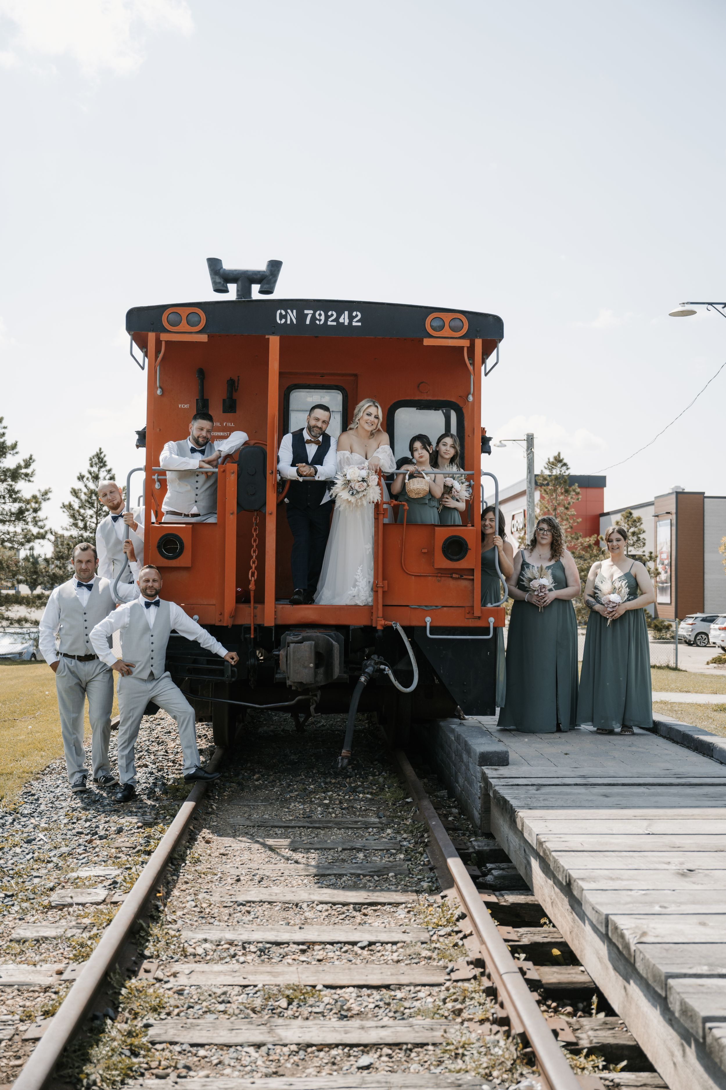 A wedding party poses with an orange vintage train caboose on railroad tracks during a unique outdoor photo session.