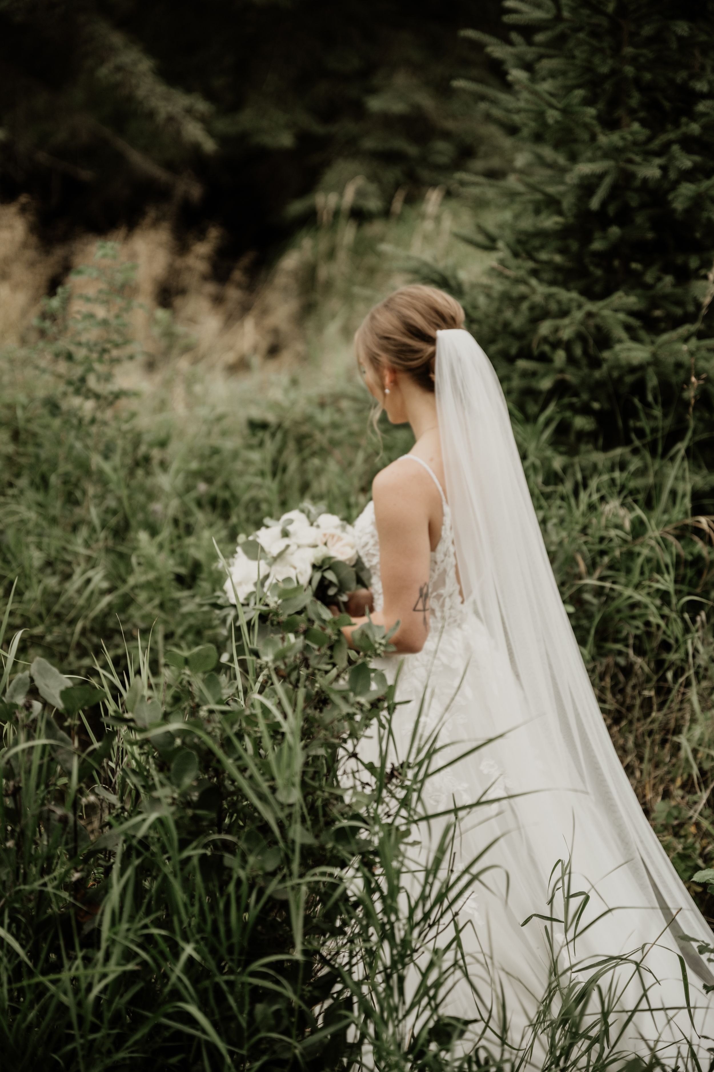 Bride in elegant white wedding dress holds bouquet while standing in lush green outdoor setting with flowing veil.