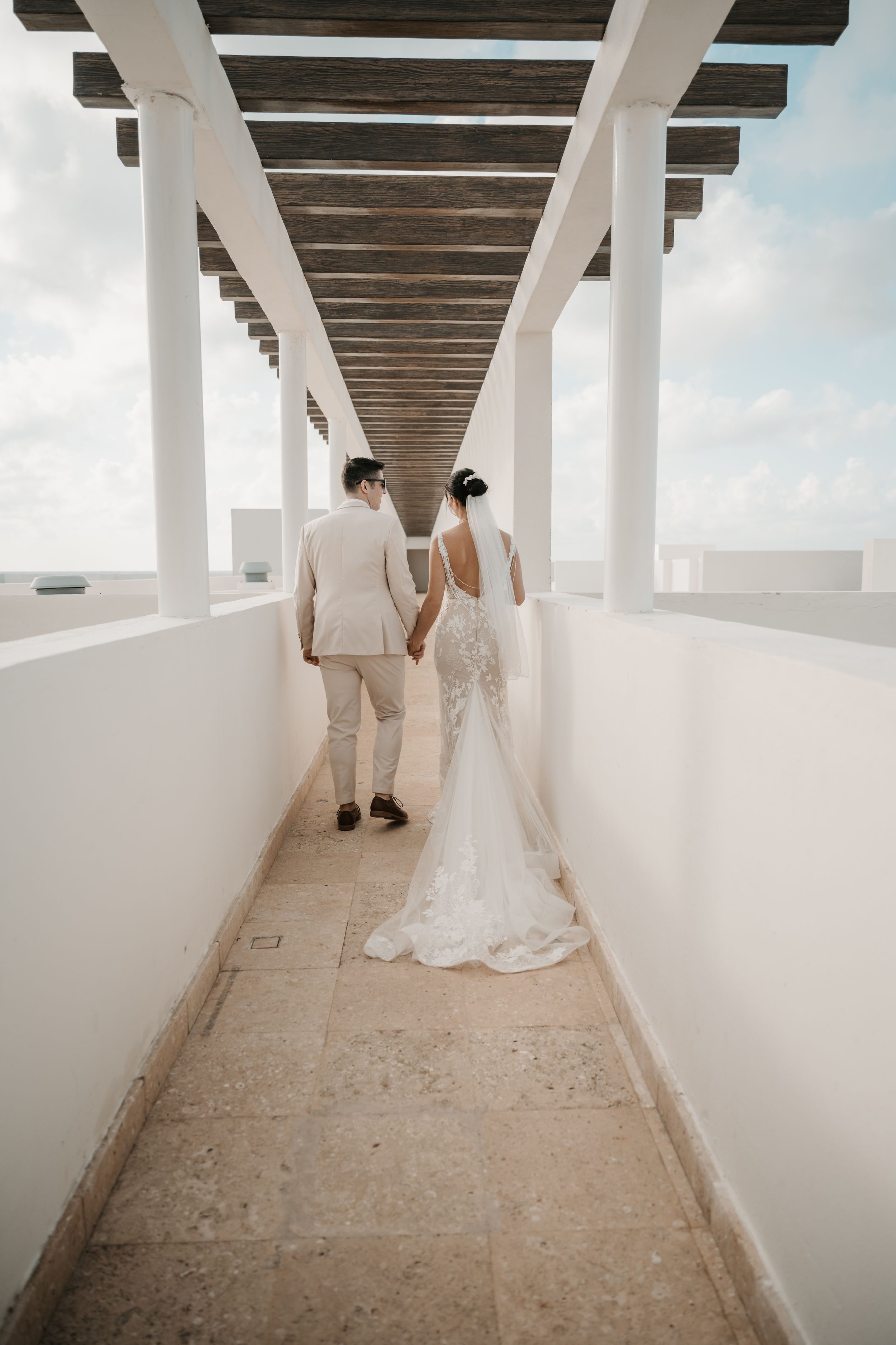 A couple in wedding attire holds hands while walking down a white covered passageway with wooden beams overhead.