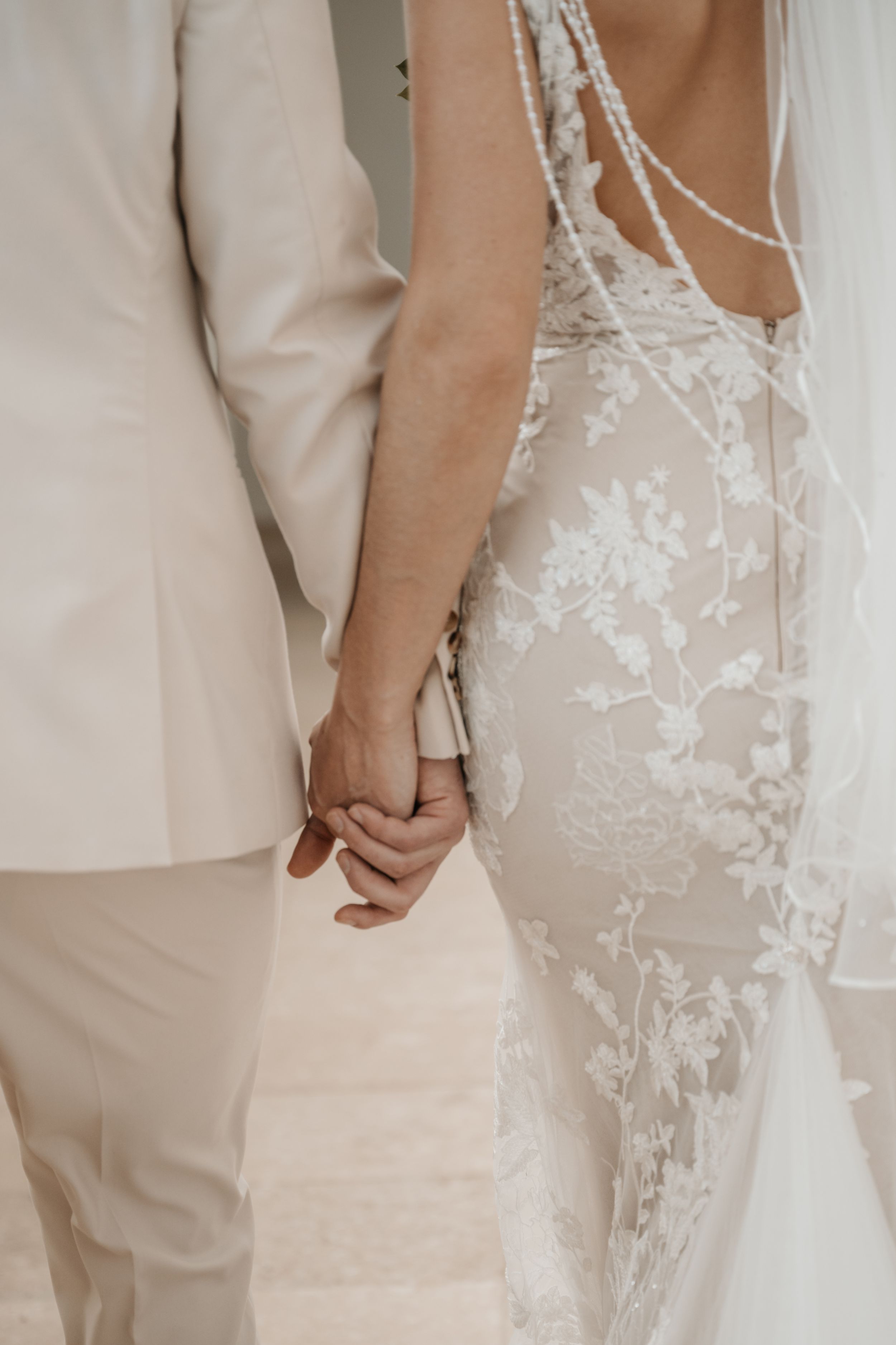 Couple holds hands at wedding ceremony wearing elegant white lace gown and light beige suit.