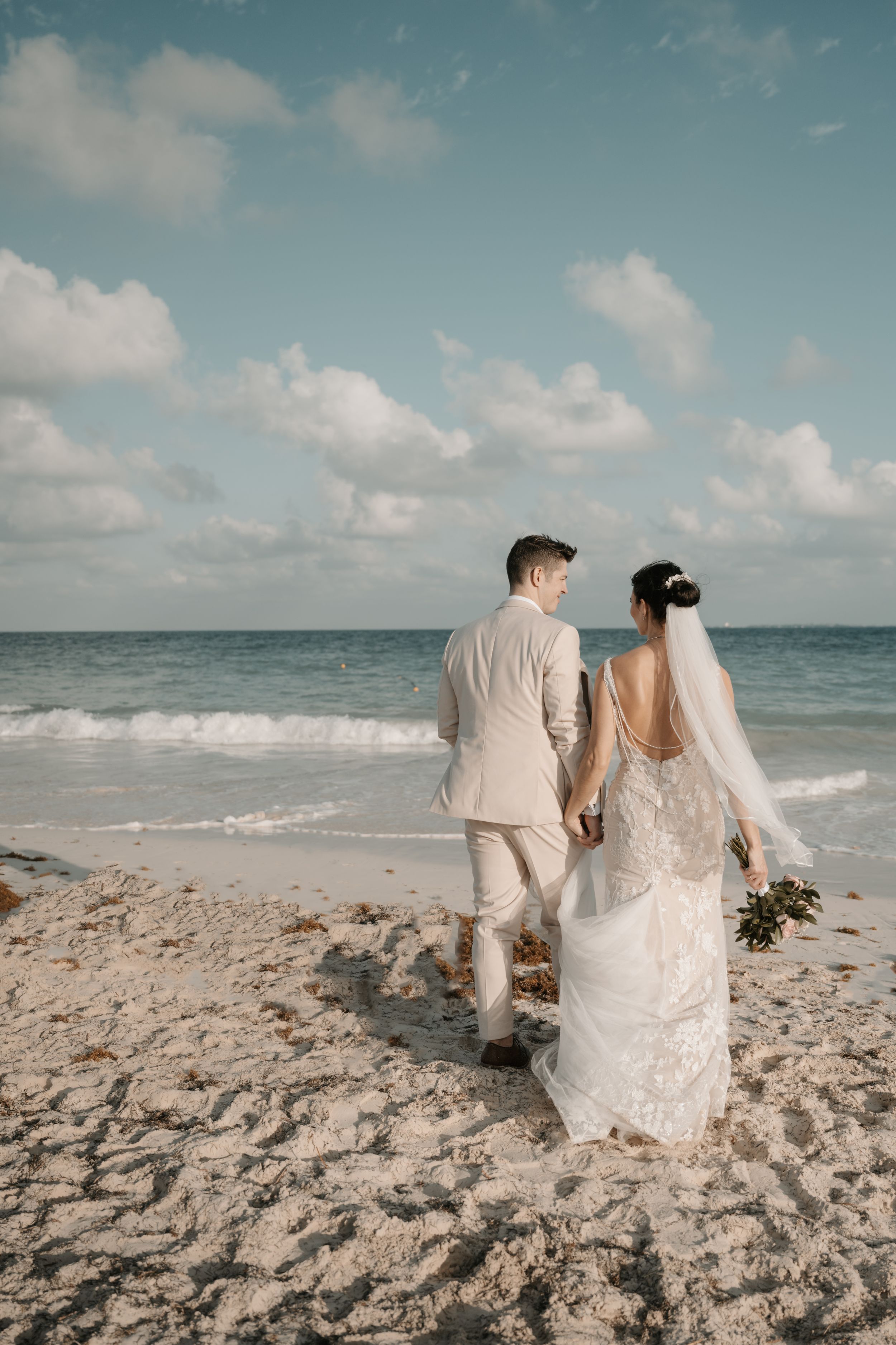 A couple holds hands while walking on a sandy beach during their seaside wedding ceremony.