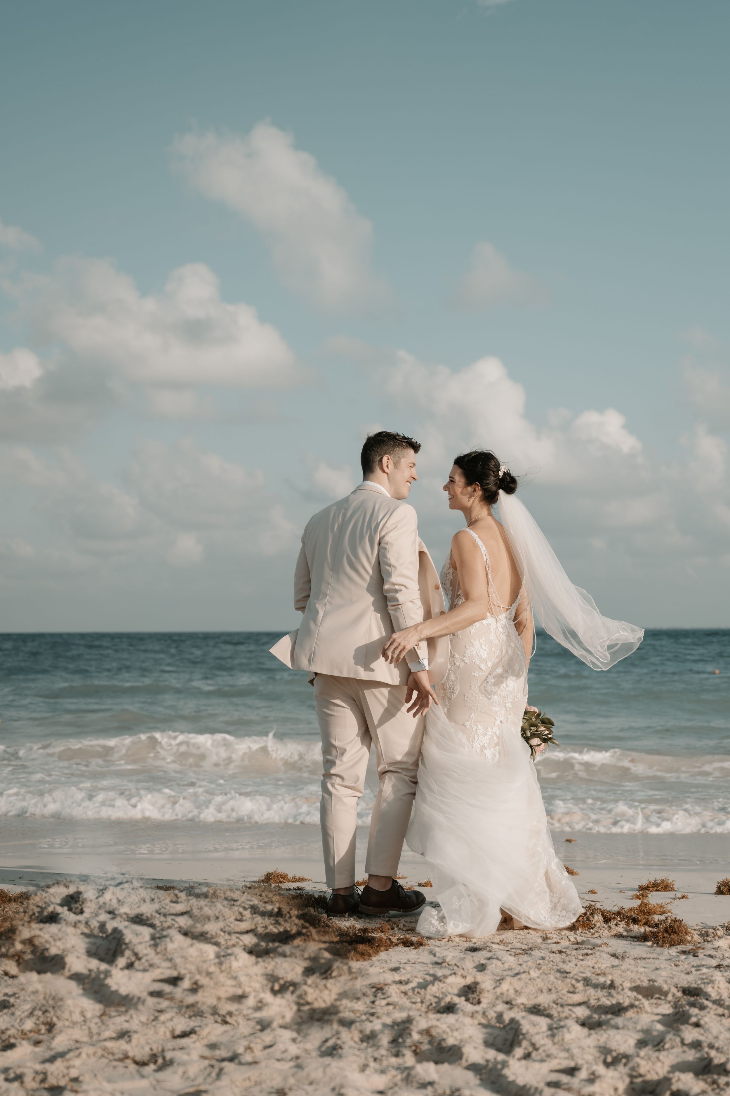 A couple in wedding attire holds hands while standing on a beach with turquoise waters and white clouds in the background.