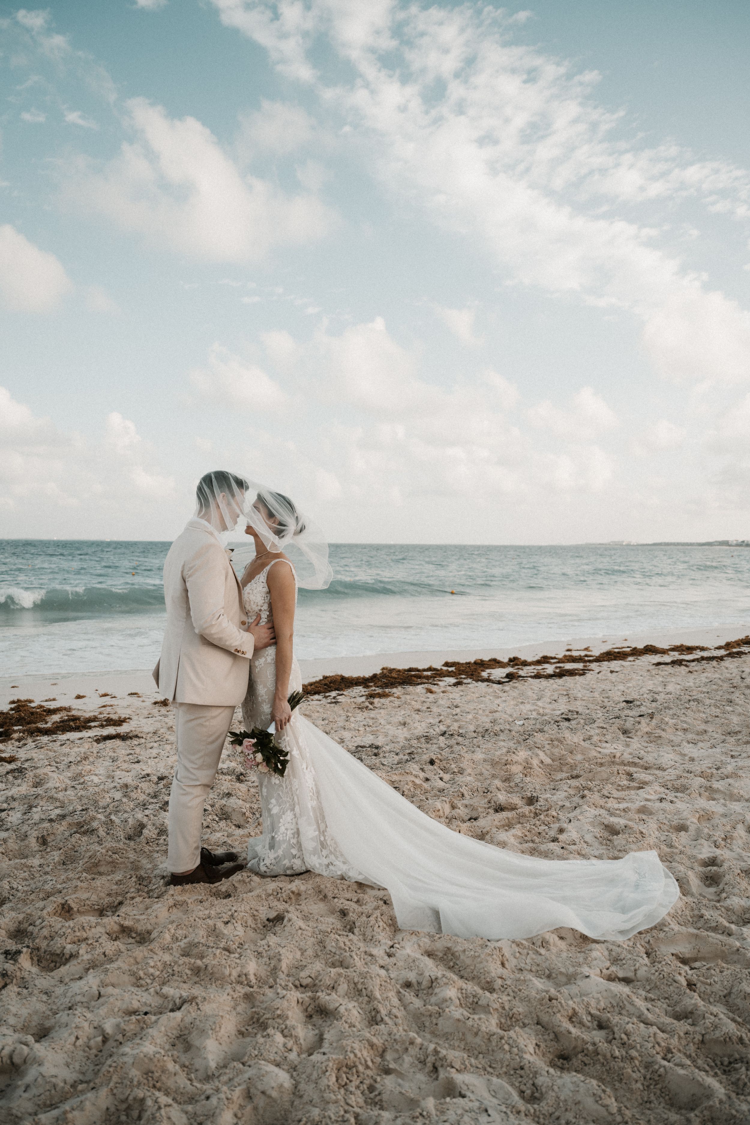 A romantic beach wedding couple embraces on sandy shores with a flowing white dress and ocean waves in the background.
