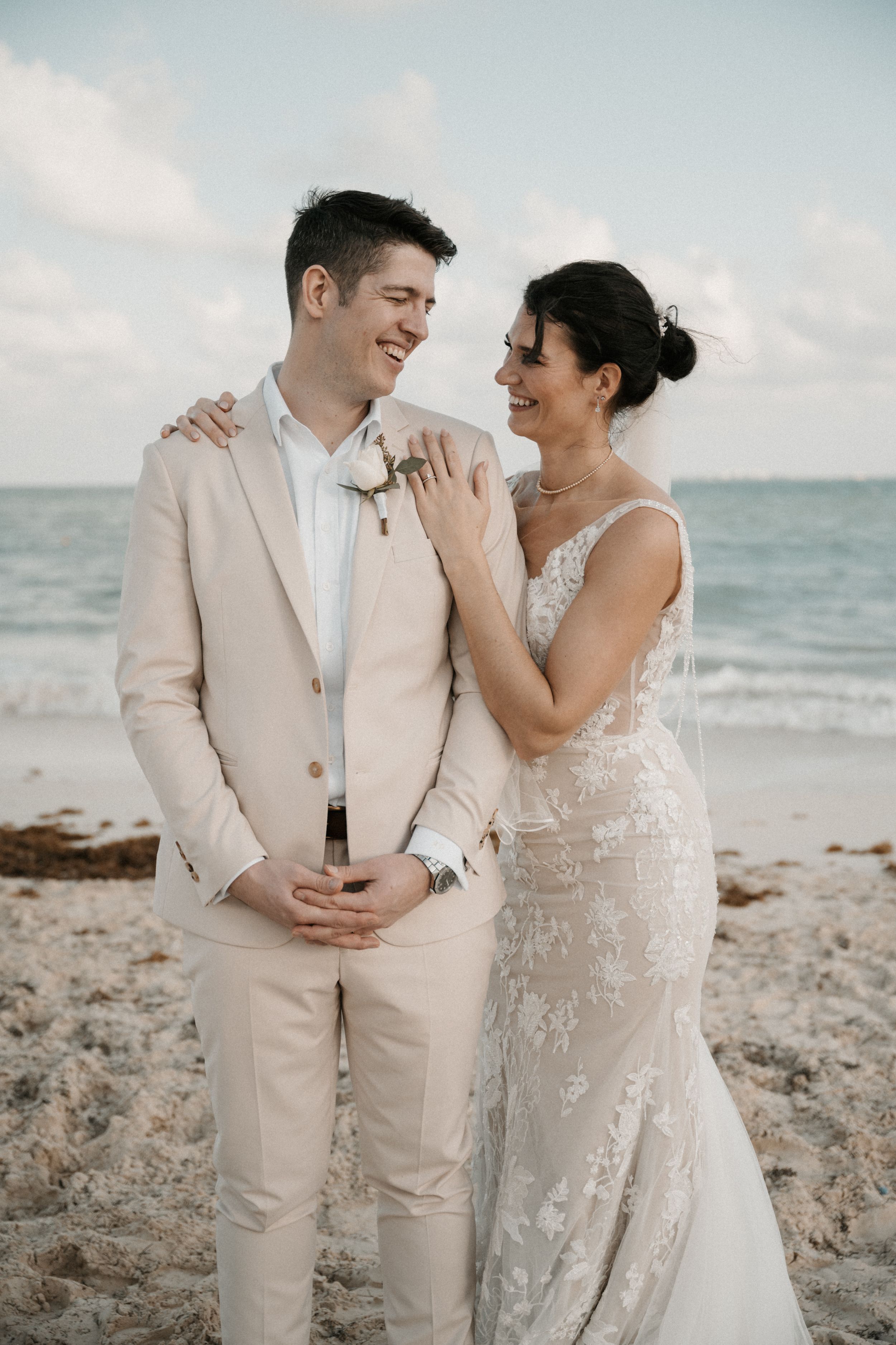 A romantic beach wedding photo of a couple in light formal attire smiling and embracing by the ocean at sunset.