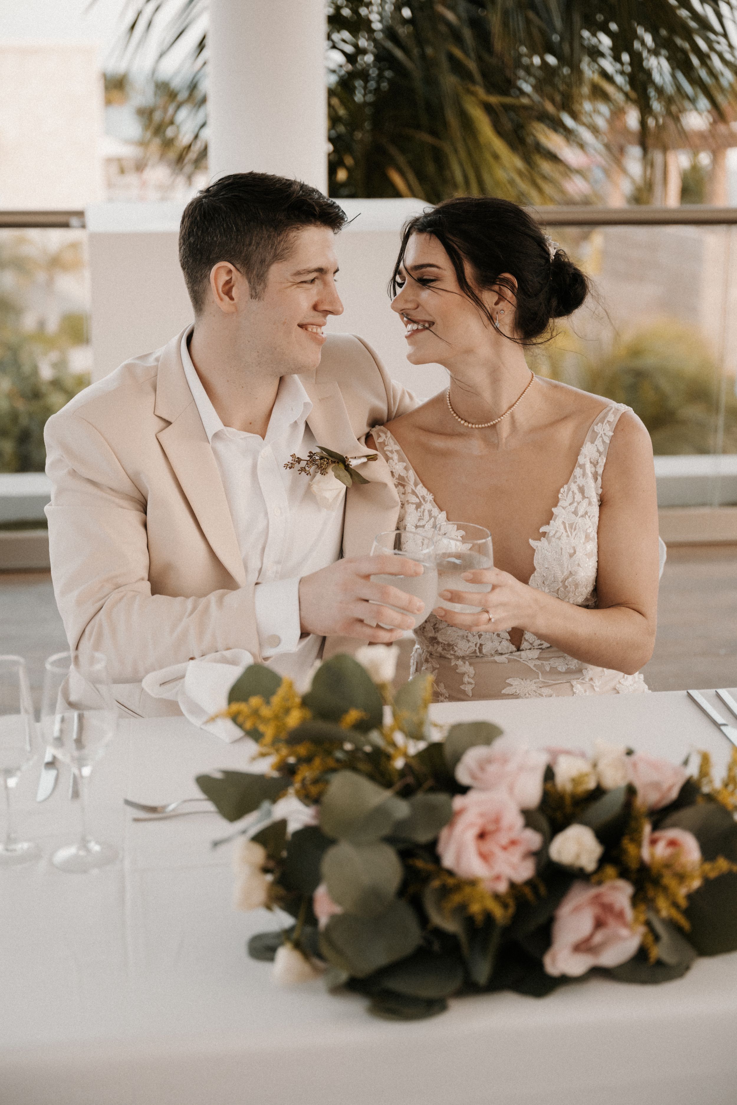 A romantic wedding couple shares a candid moment at their reception table with blush floral centerpiece and palm trees in background.