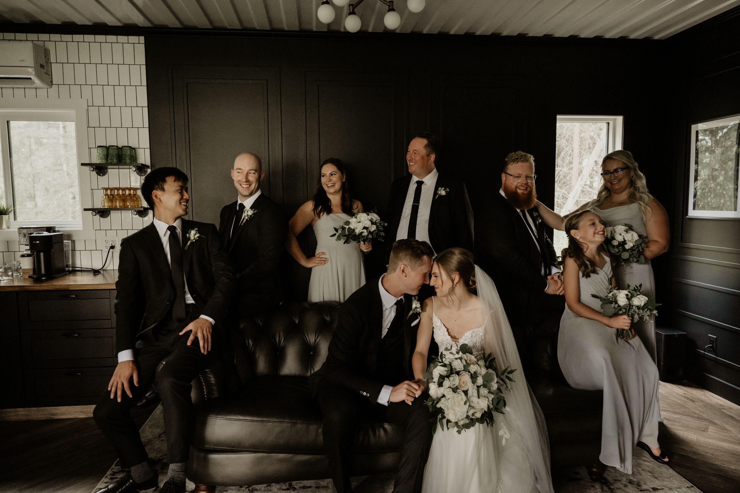 Wedding party in formal attire gathered in a rustic indoor venue with dark walls and white trim.