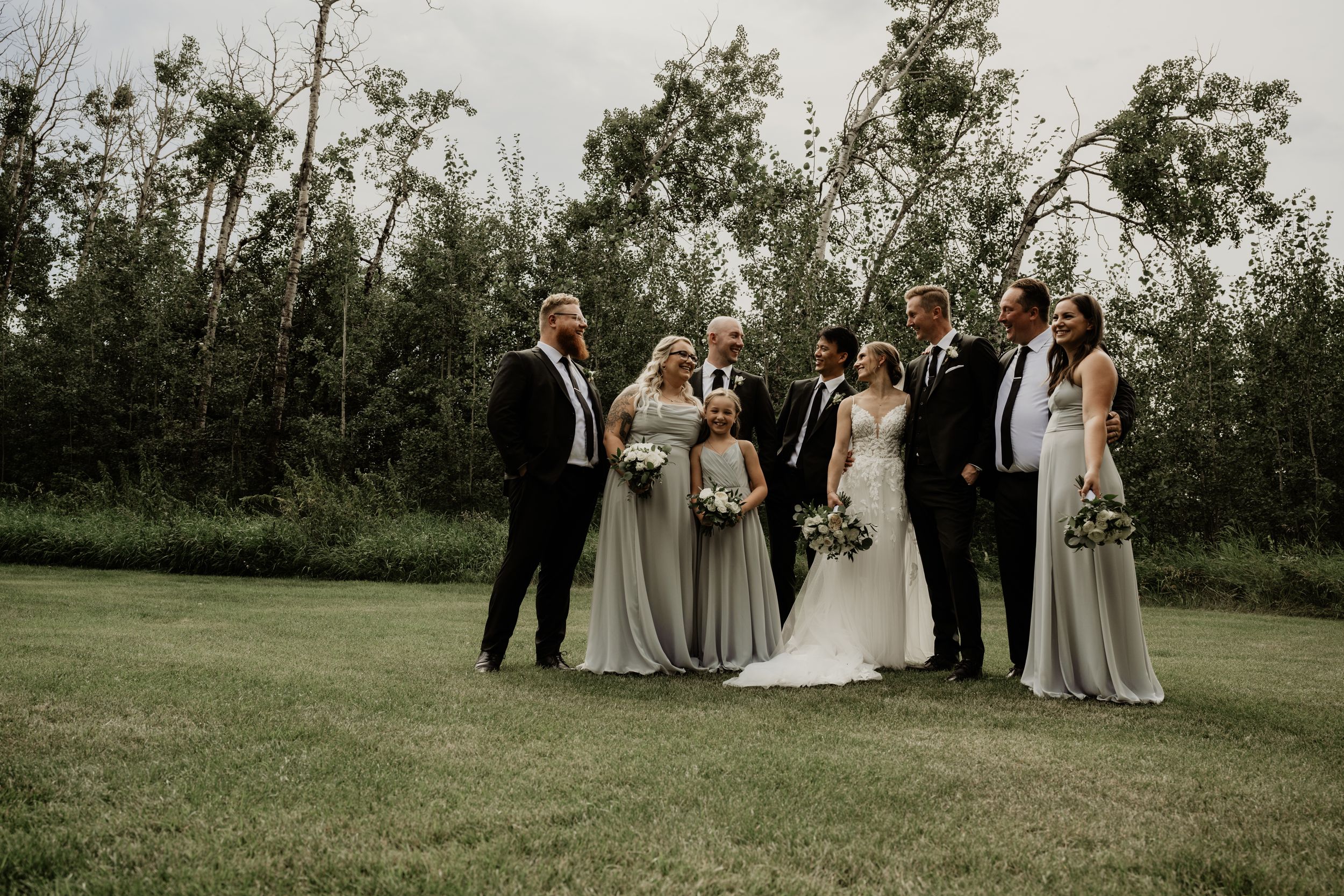 Wedding party in formal attire stands together on a green lawn with trees in the background at dusk.