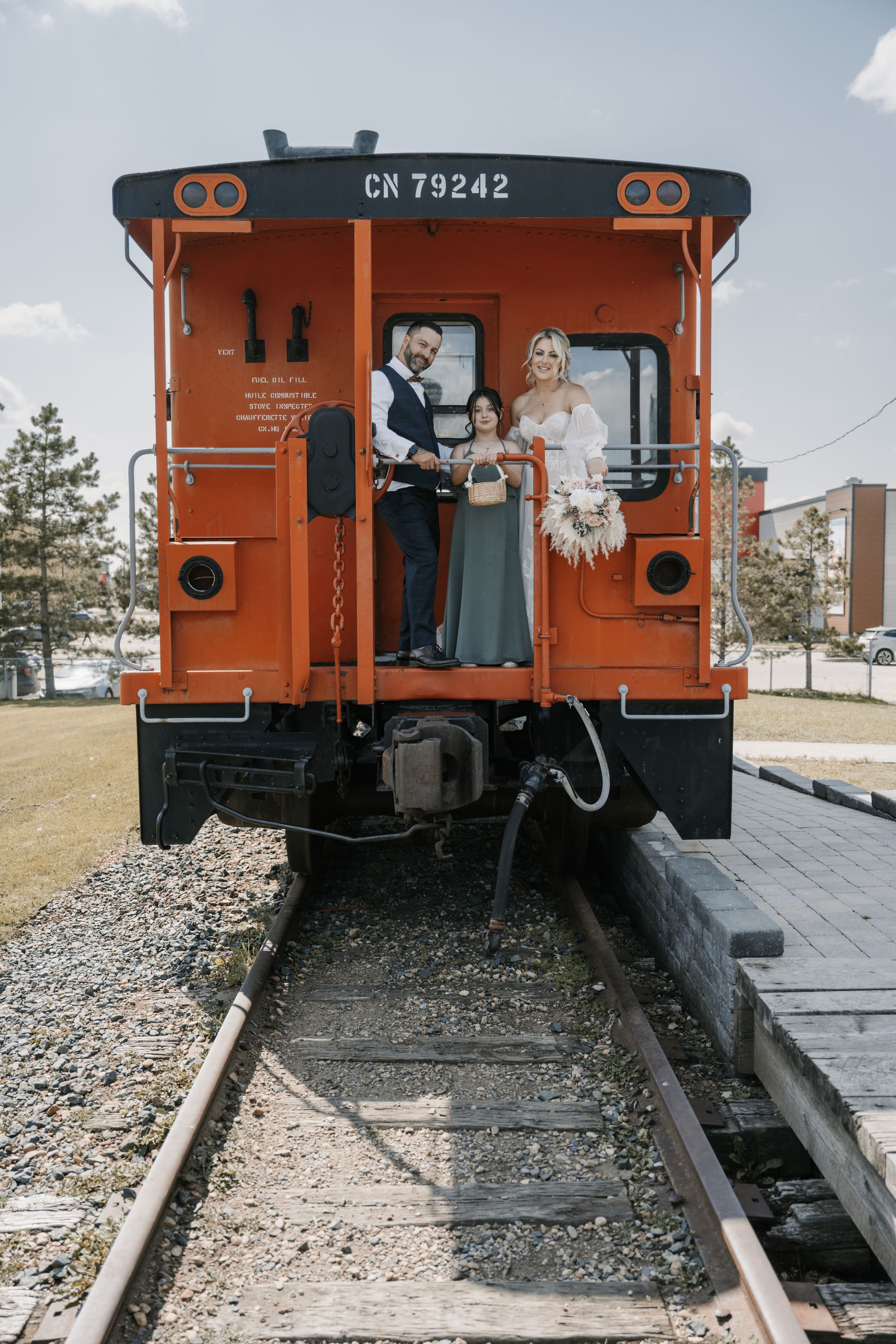 A couple stands at the back entrance of a vintage orange CN 79242 train car on railroad tracks.