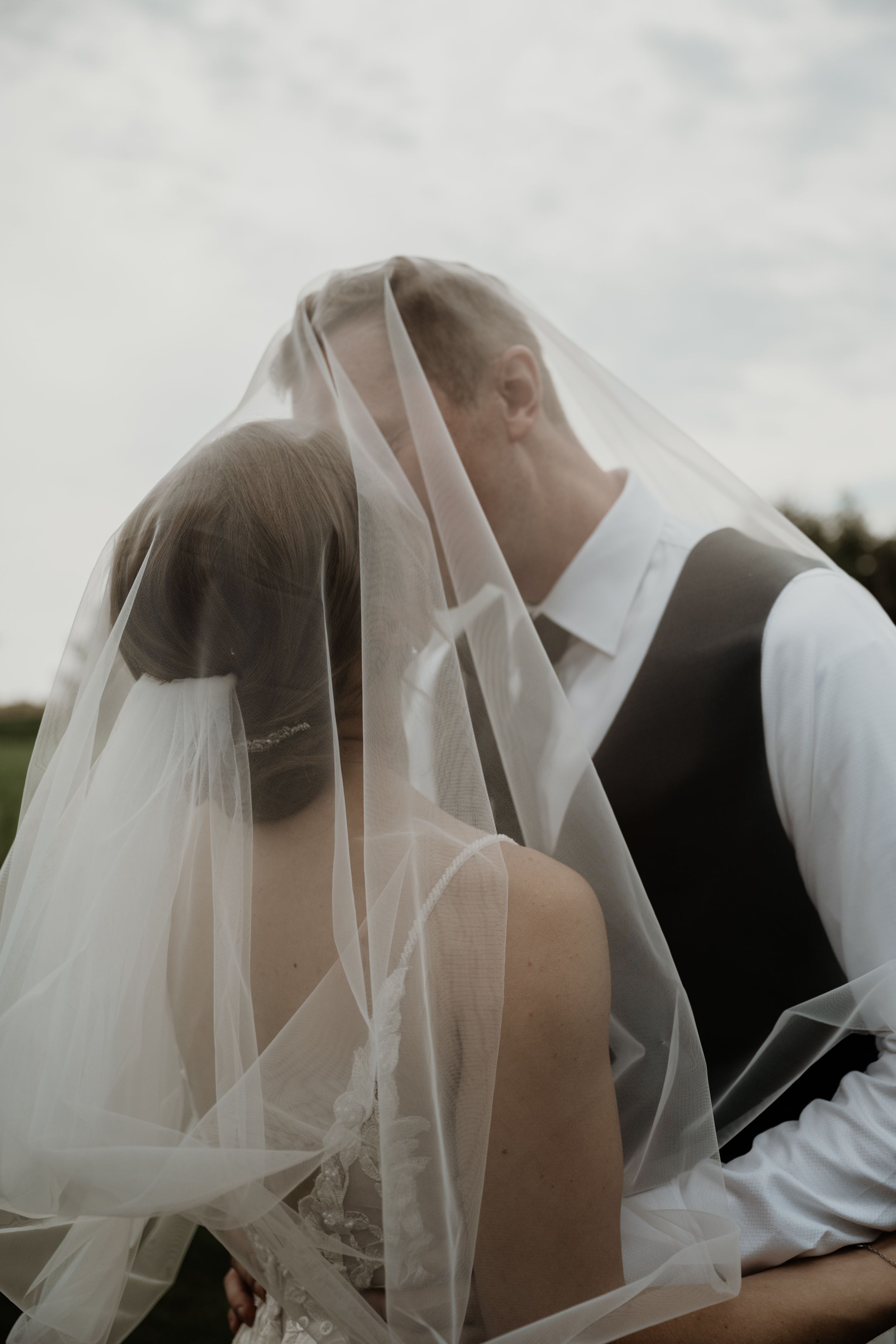 Intimate wedding photography showing couple embracing under delicate sheer veil outdoors in natural lighting.