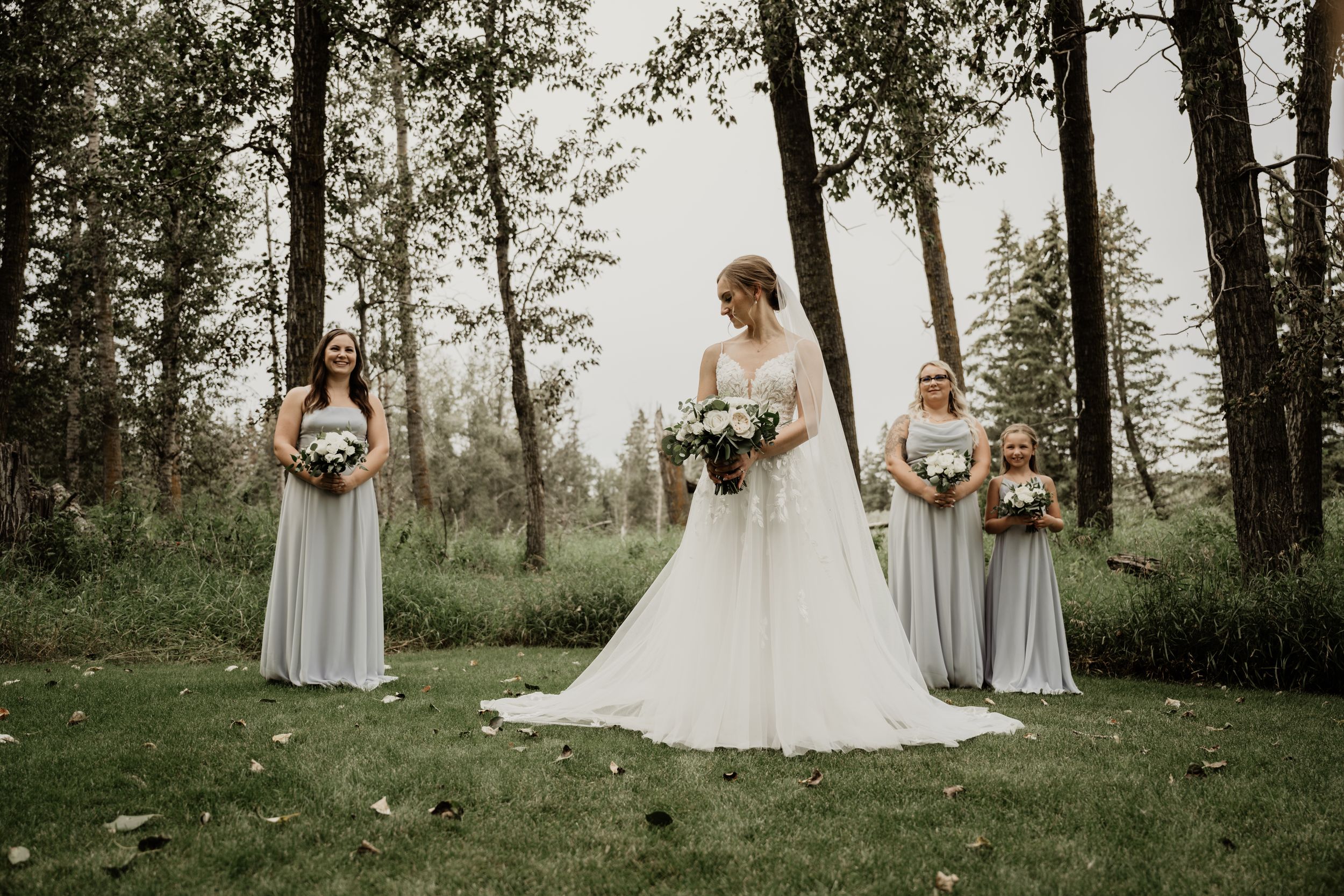 A bridal party in light gray dresses and white gown poses elegantly among tall trees in a dreamy forest setting.