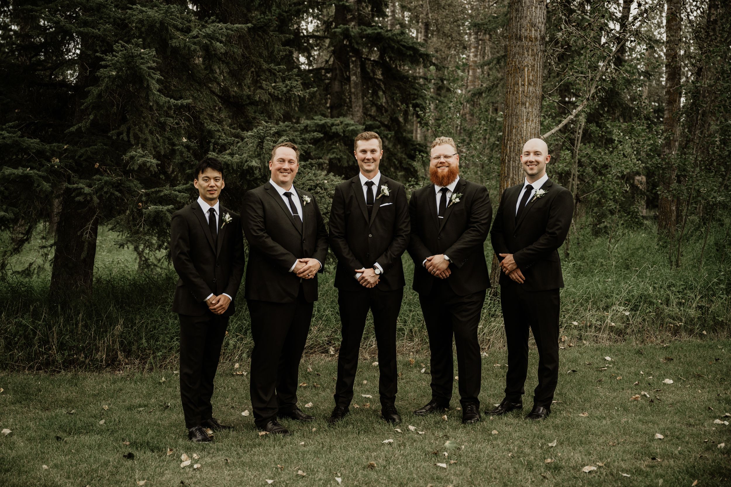 A wedding party group of five people wearing black suits and ties pose together in a lush forest setting.