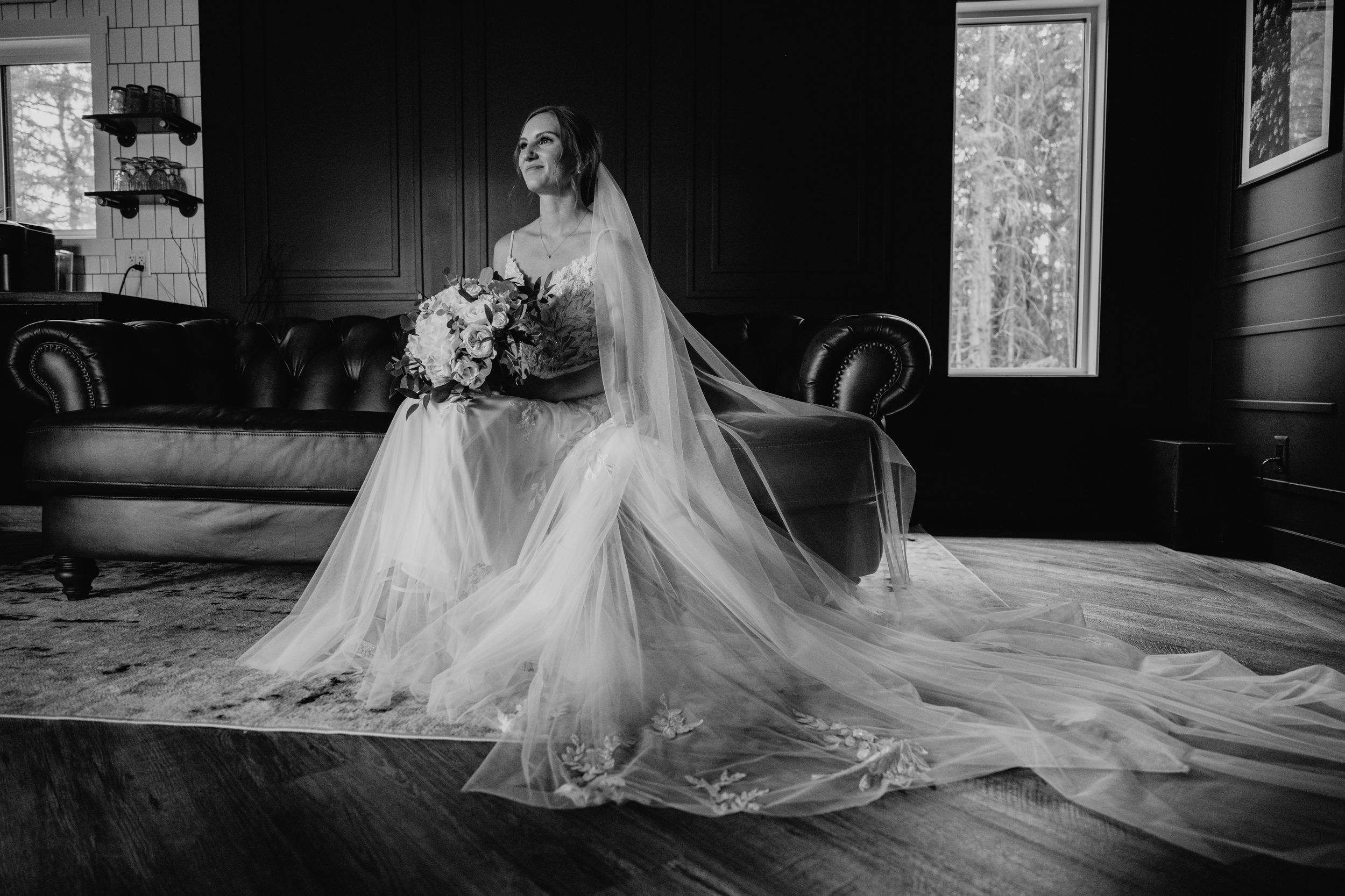 A stunning black and white wedding photograph showing a figure in a flowing lace gown and veil seated on an elegant sofa.