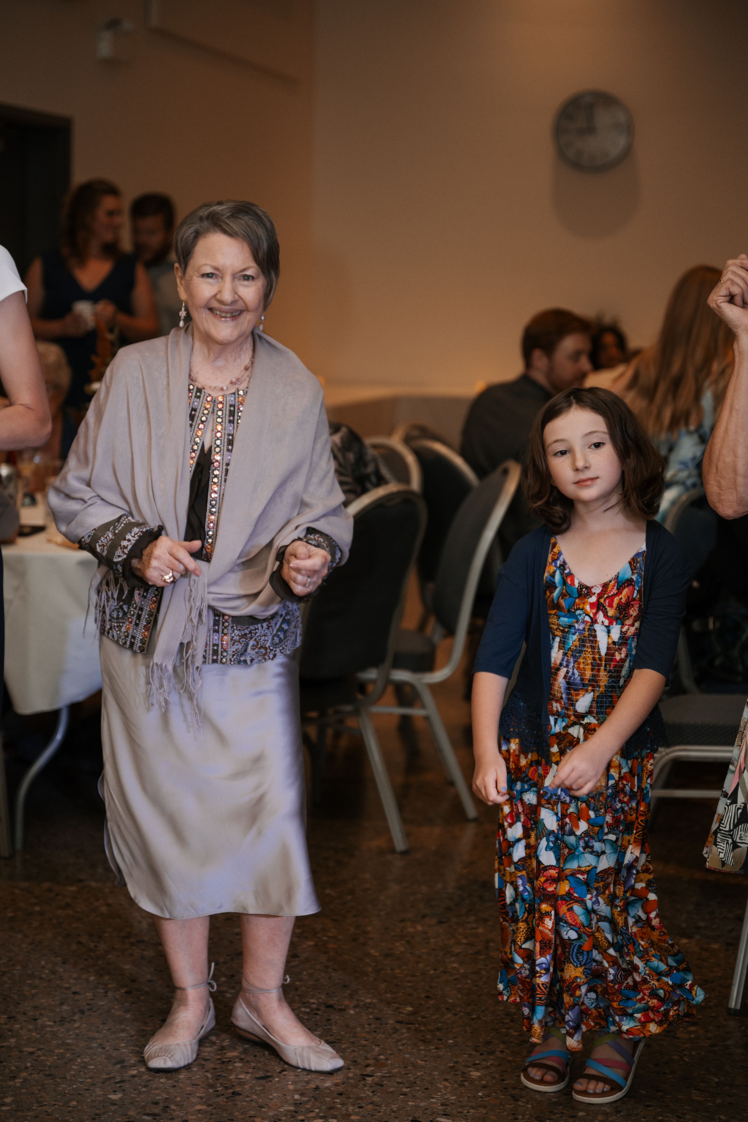An elderly woman in grey cardigan and a young girl in a colorful floral dress are standing together at an indoor event.