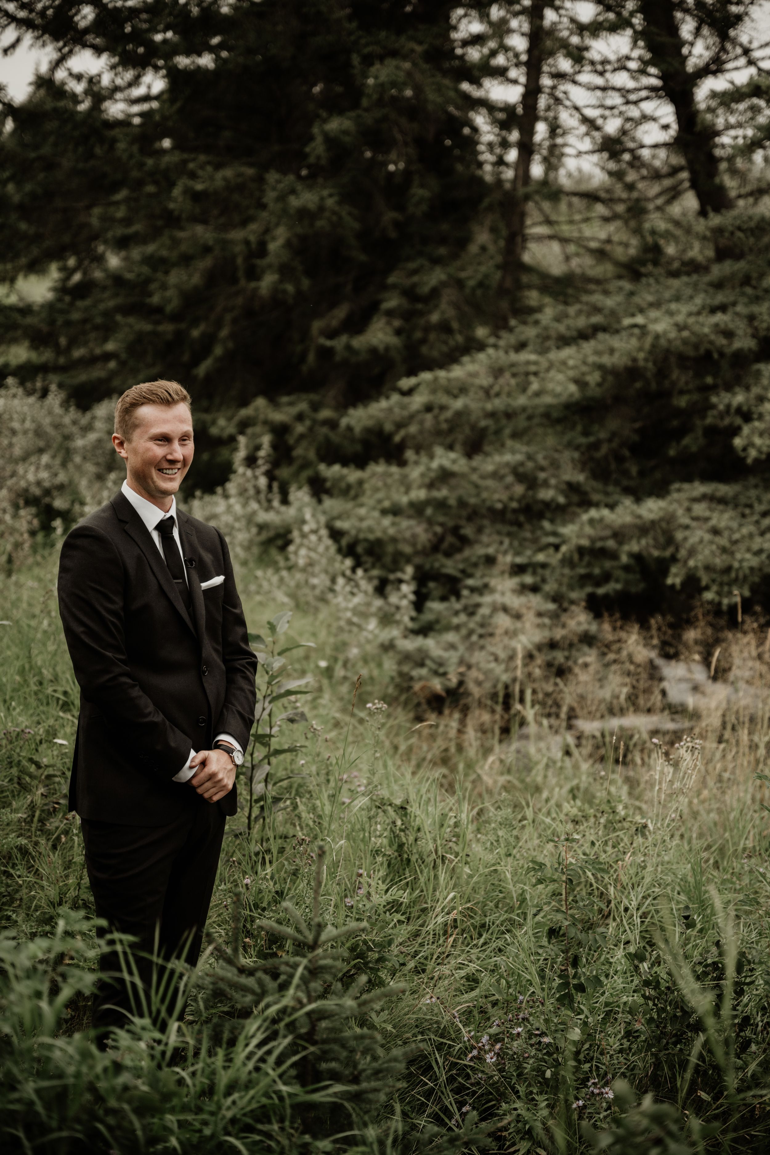 Person in a black formal suit stands smiling in tall grass with evergreen trees in the background on an overcast day.
