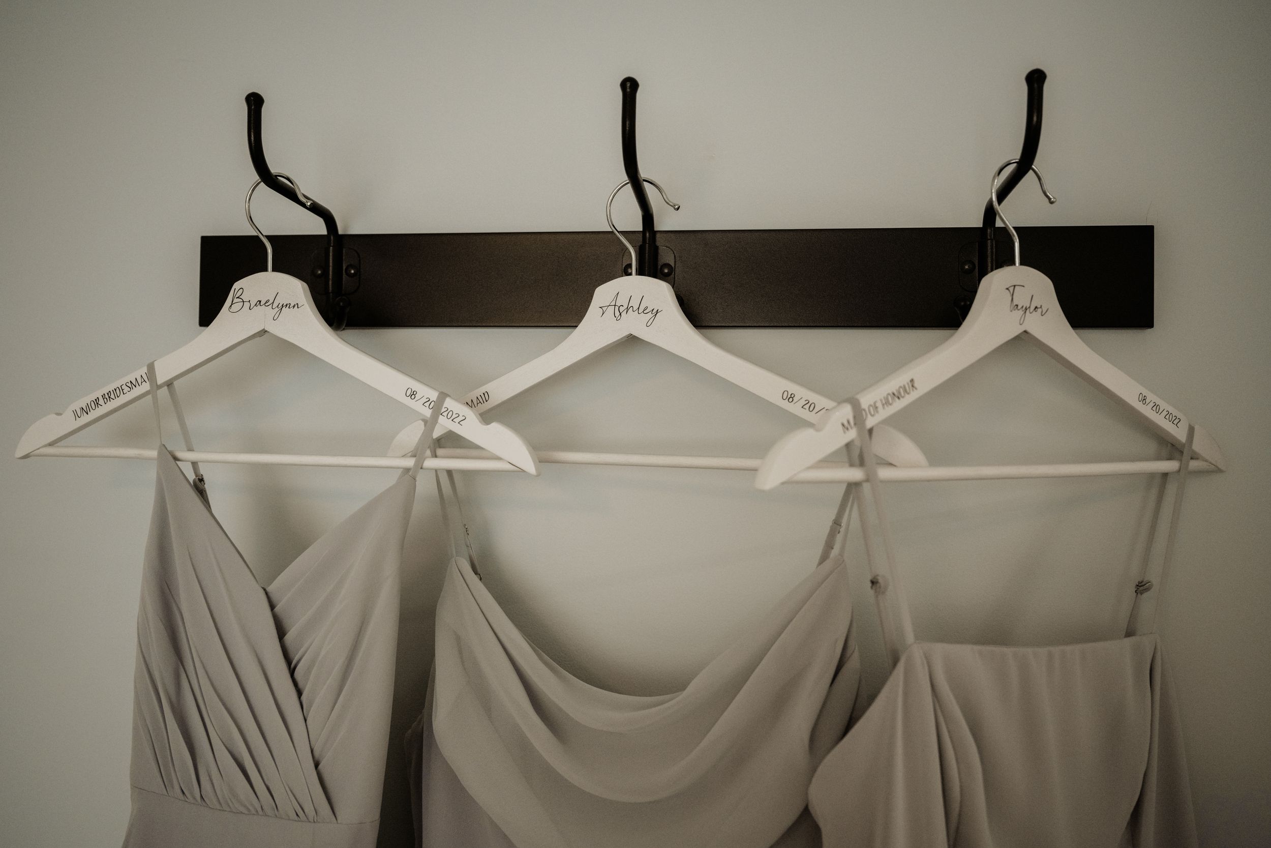 Three elegant neutral-colored bridesmaid dresses hanging on white hangers against a dark wooden wall-mounted coat rack.
