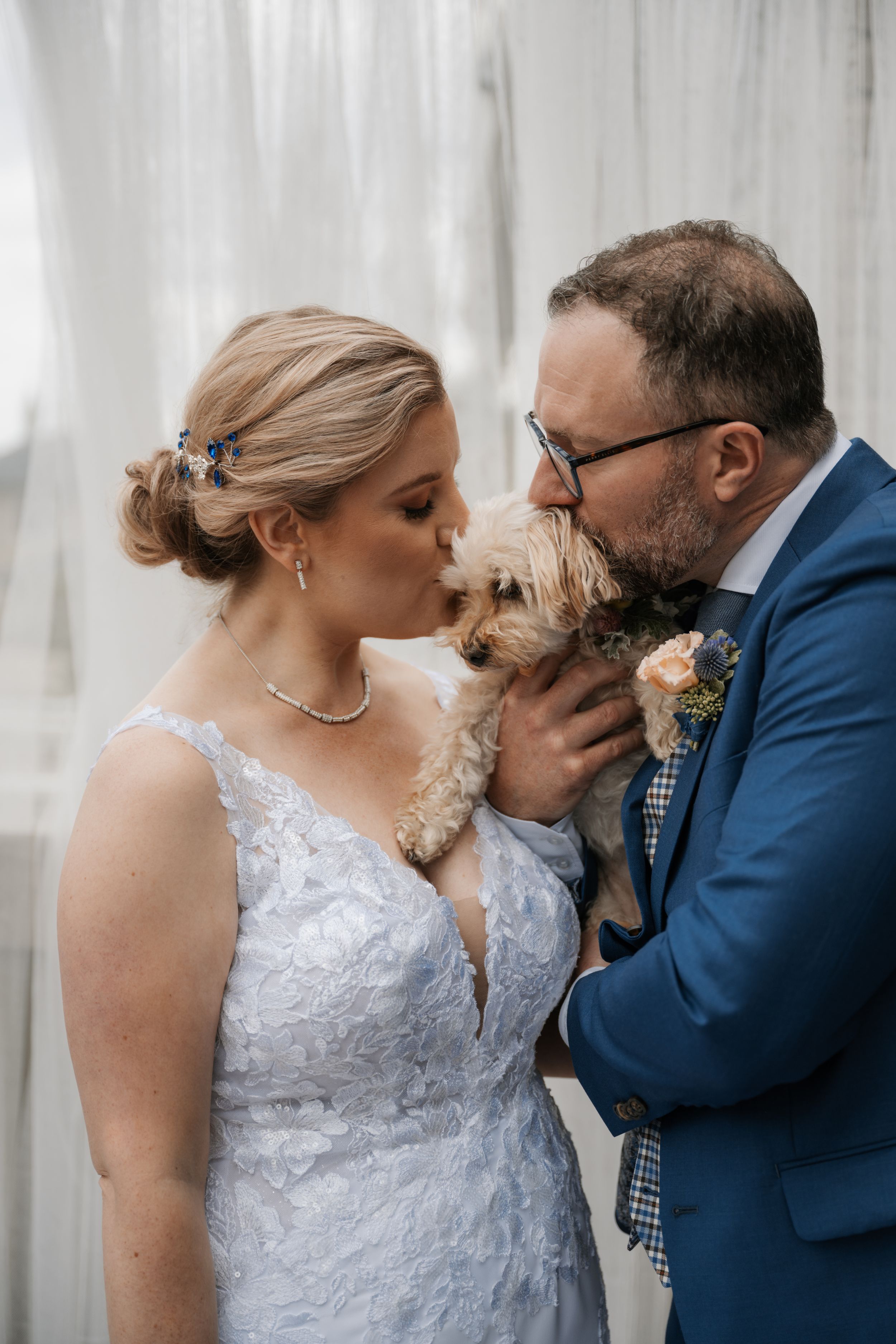 A couple in wedding attire sharing a tender moment while holding their small terrier dog at their wedding celebration.