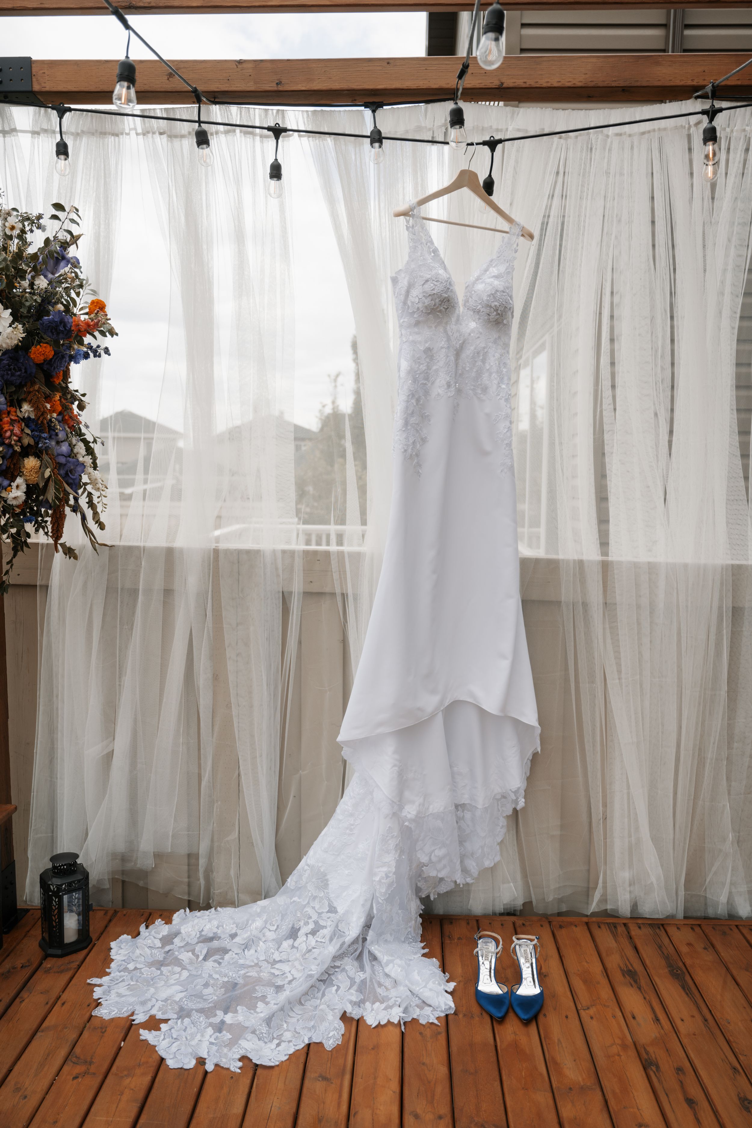 A white lace wedding gown with dramatic train hangs on display against sheer curtains, with blue heels placed beneath.