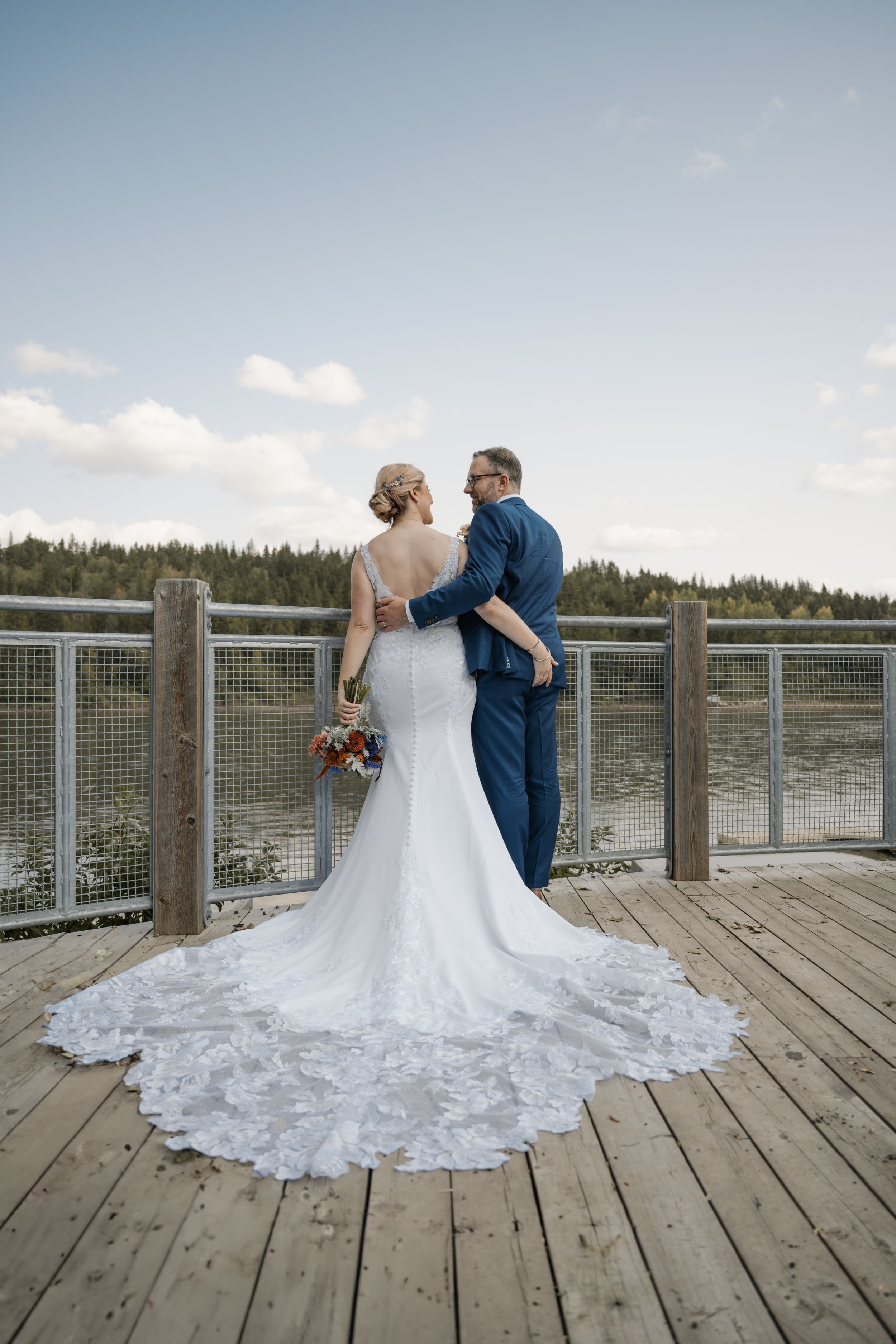 A couple in wedding attire stands on a wooden dock overlooking a lake, with the bride's lace dress train flowing behind them.